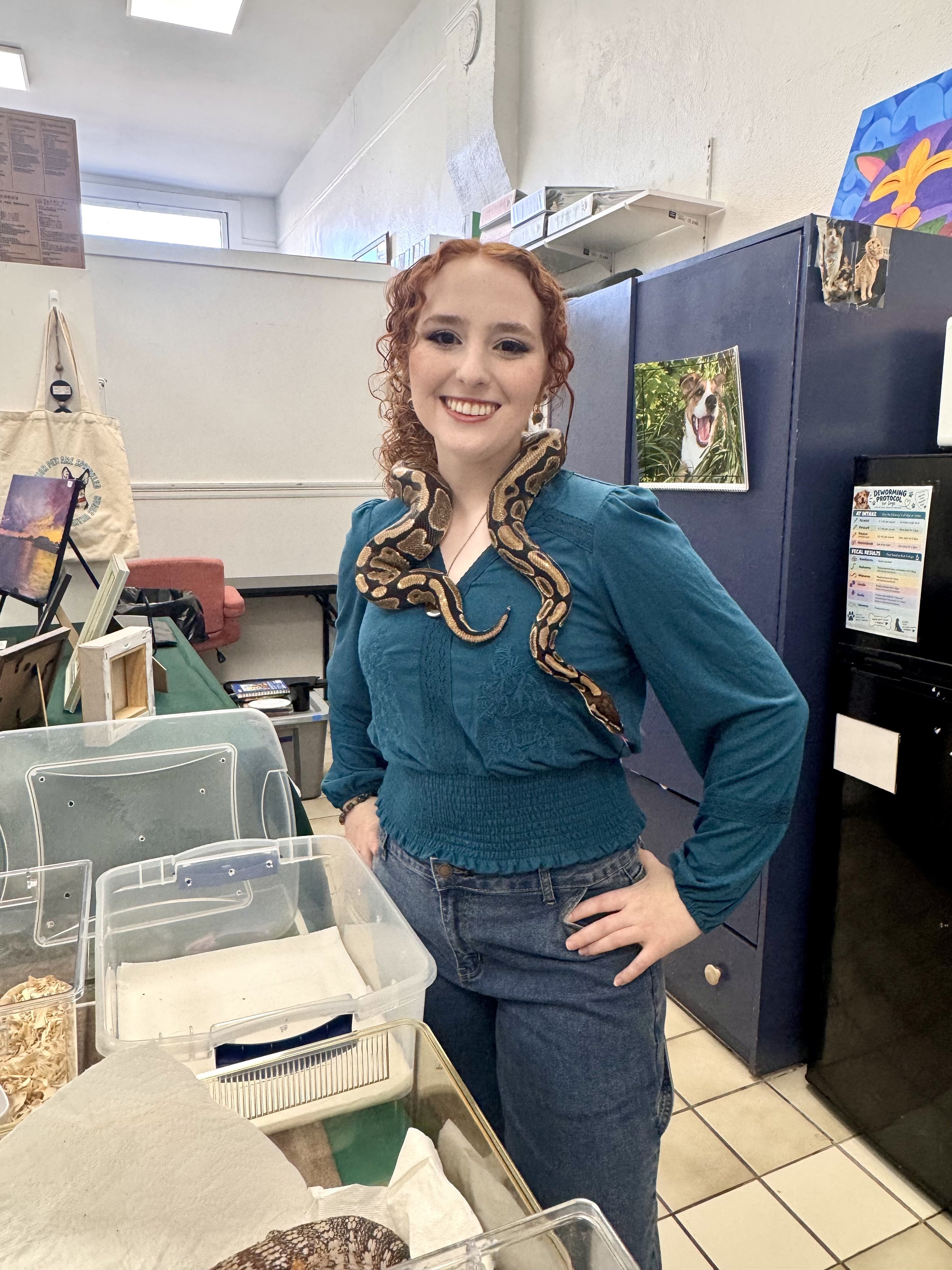 A woman with curly red hair and makeup, wearing a teal top and jeans, standing in a room with a snake draped over her shoulders. She is smiling and posing for the camera.