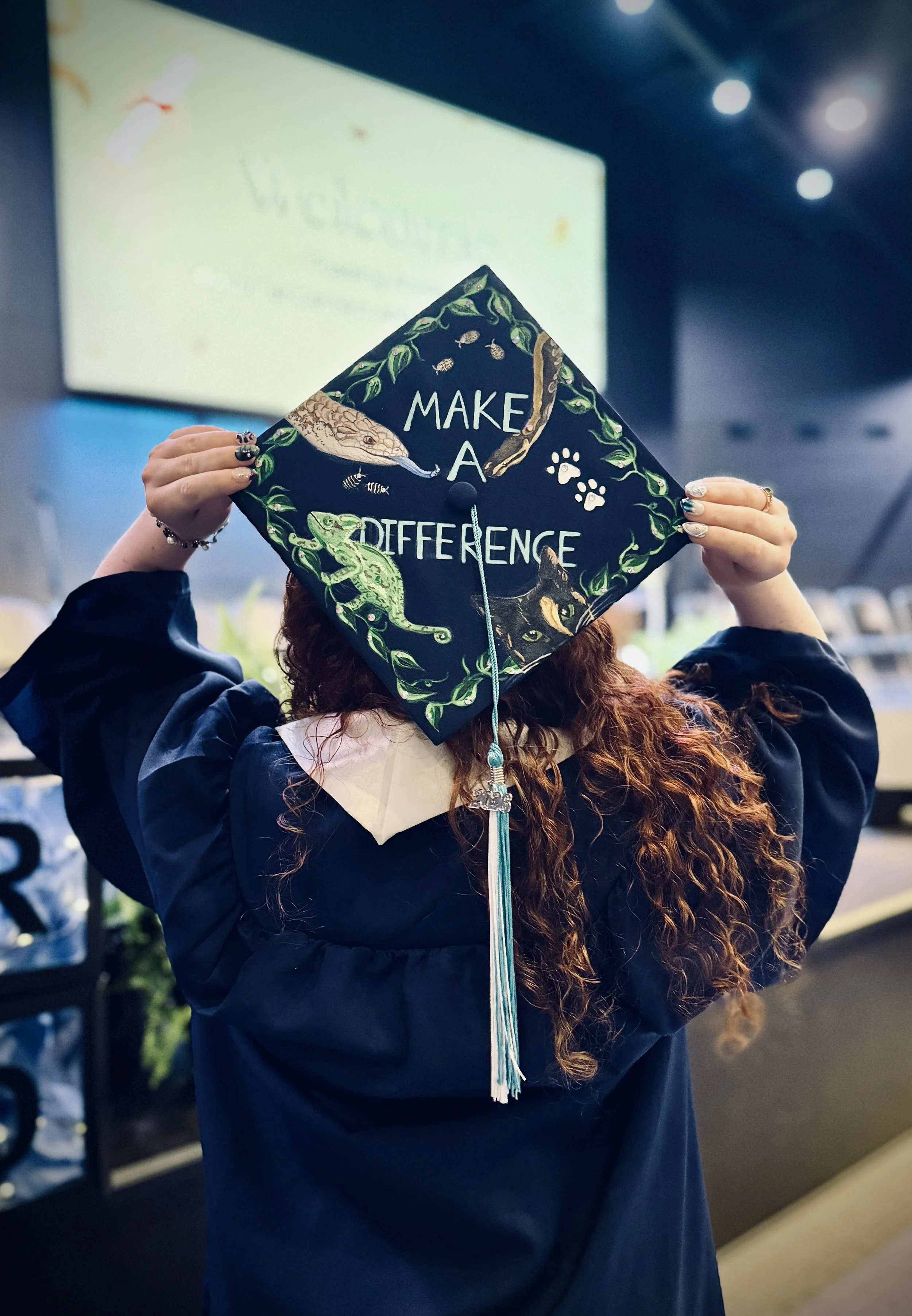 Person wearing a navy graduation gown holding a decorated graduation cap with a snake and the words 'Make a Difference' in a classroom or auditorium.