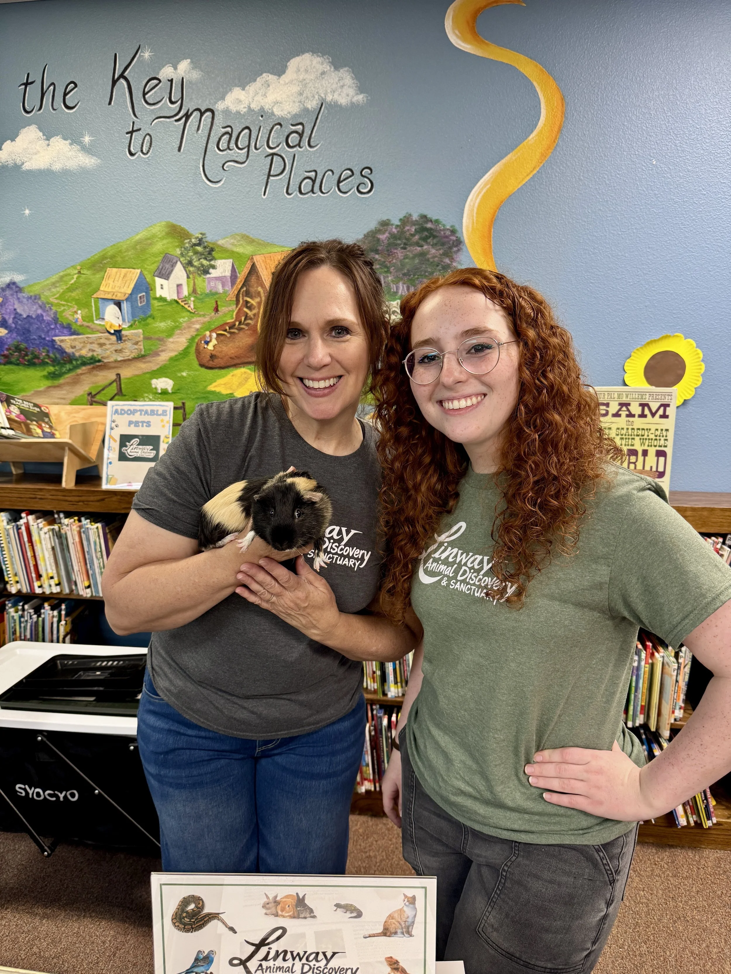 Two women standing together in a library, one holding a guinea pig. A colorful mural with clouds, houses, and a yellow spiral in the background. A sign in front promotes Linway Animal Discovery.