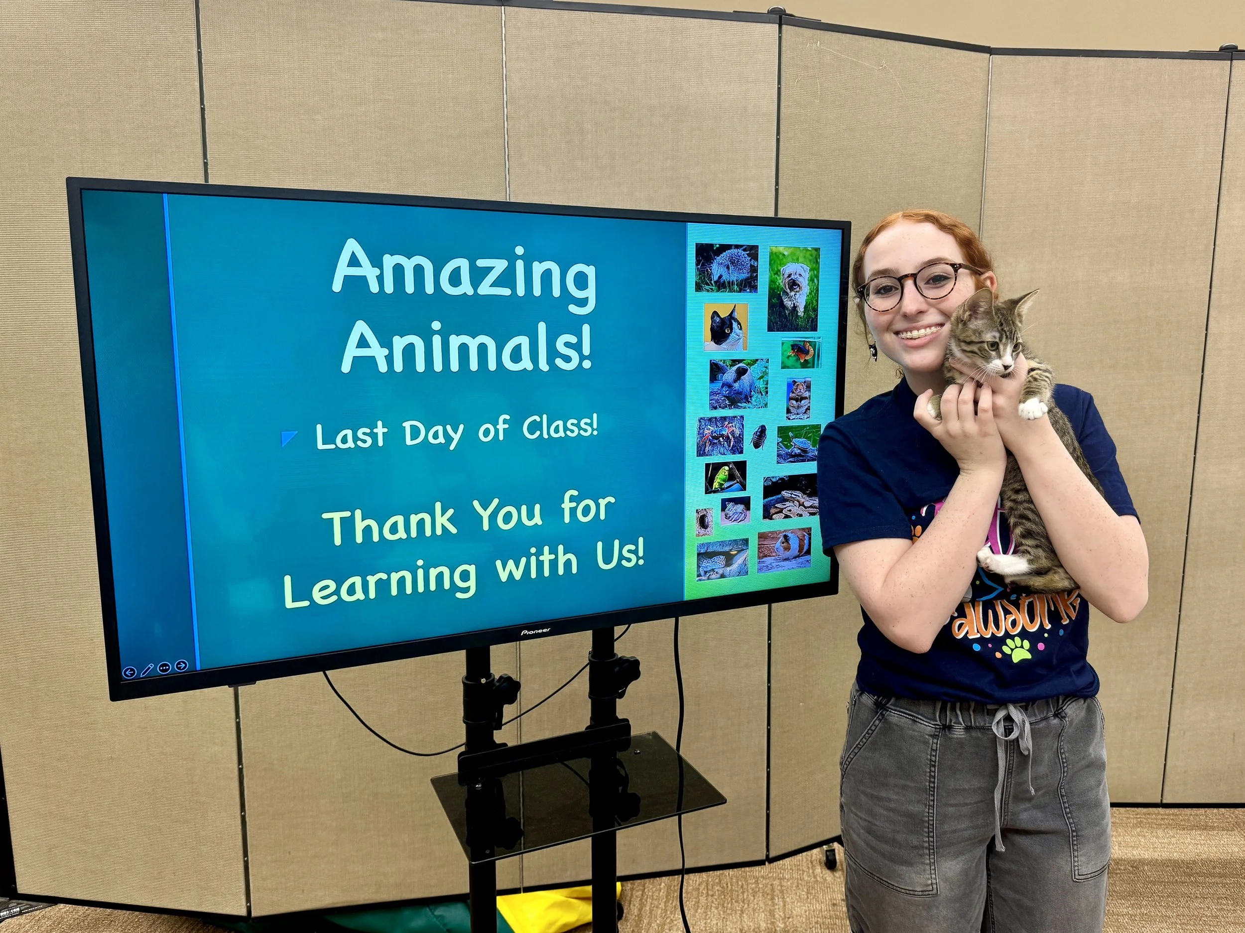A young girl with glasses is holding a tabby cat and smiling. Behind her is a large screen with a blue background displaying the words 'Amazing Animals! Last Day of Class! Thank You for Learning with Us!' and several pictures of animals.