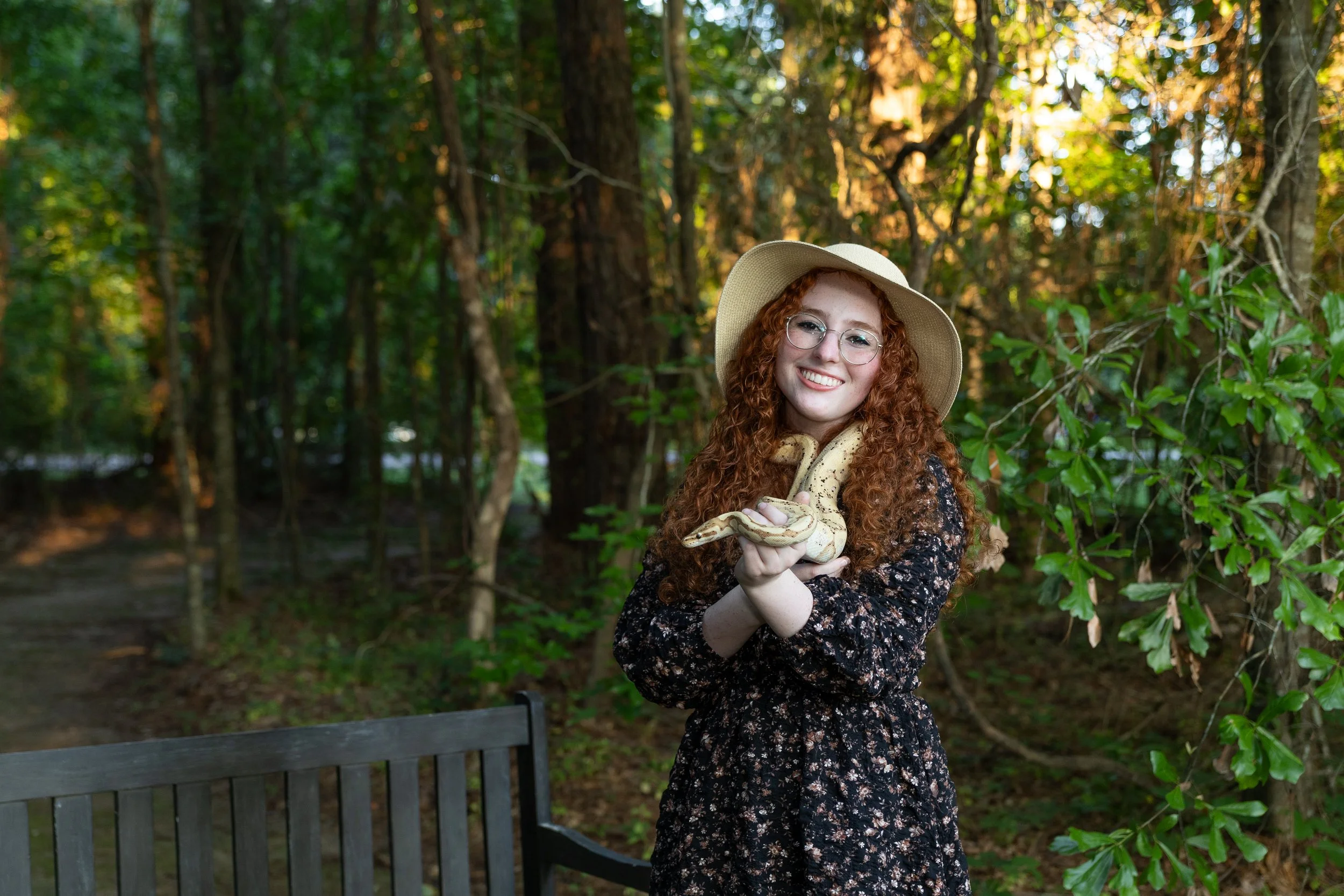 A woman with curly red hair, glasses, and a wide-brimmed hat outdoors in a wooded area, holding a snake in her hands and smiling.