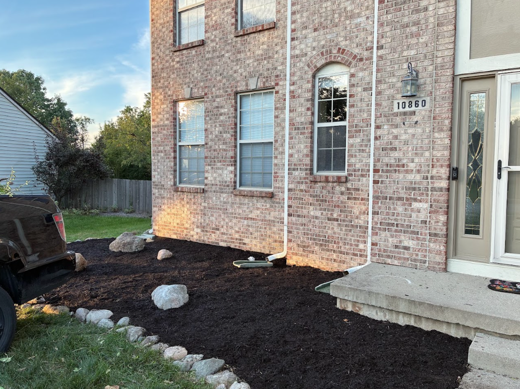 A brick house with a porch and a yard under renovation, with fresh soil and rocks along the edge, and a black truck parked nearby.