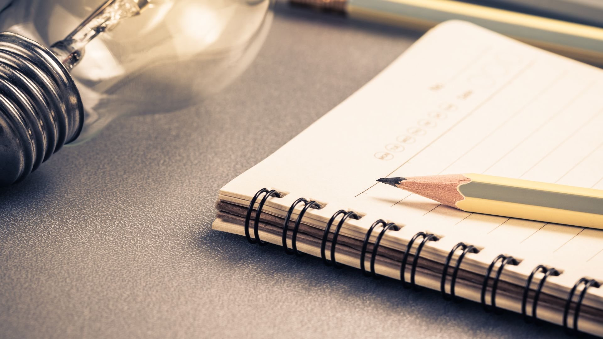 Close-up of a notepad with a spiral binding, a sharpened yellow pencil resting on the notepad, and a metallic dumbbell in the background on a gray surface.