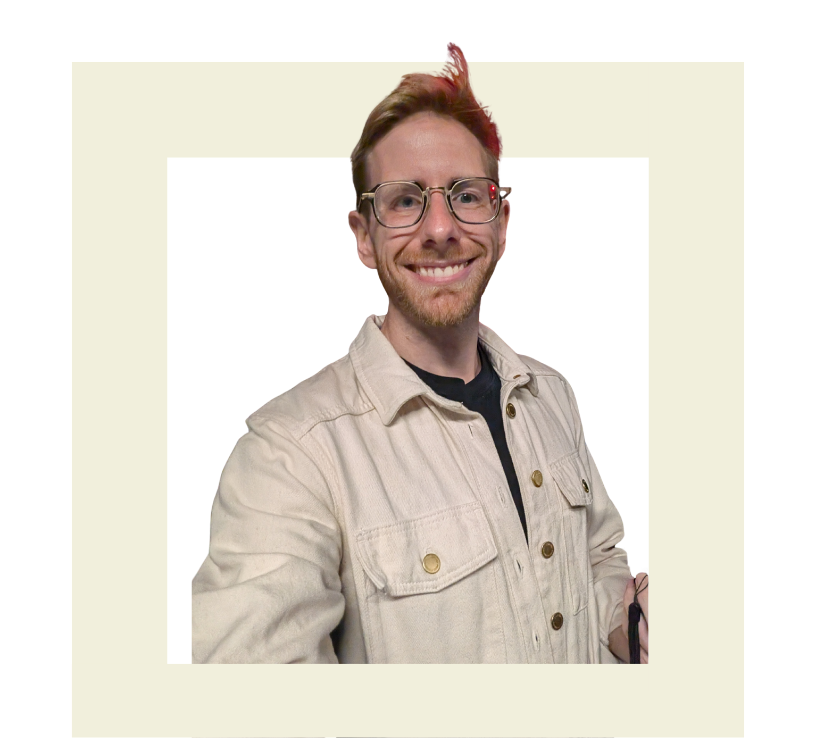 A young man with red hair, beard, and glasses, smiling and wearing a beige jacket over a black shirt.
