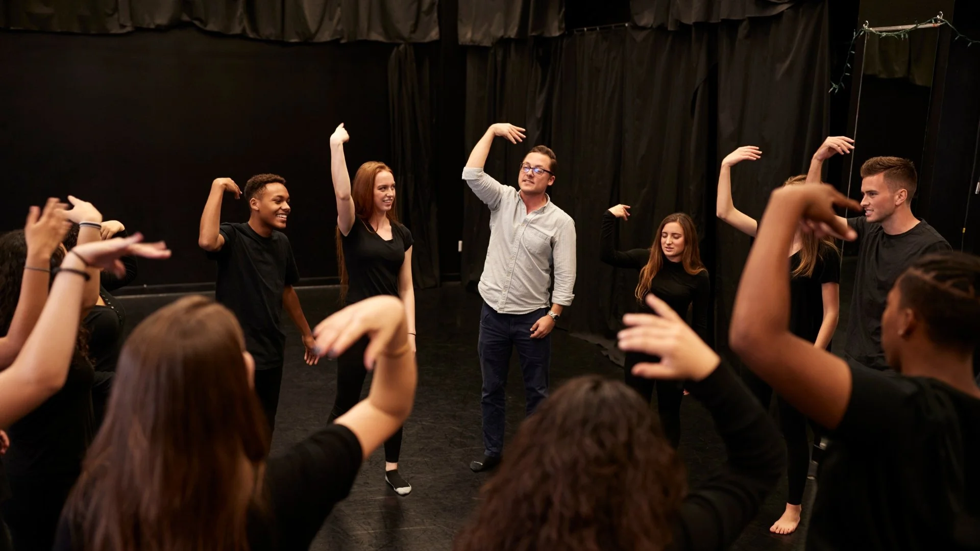 A group of diverse young adults participating in a drama or acting class, standing in a circle with their arms raised and smiling, in a black room with curtains.