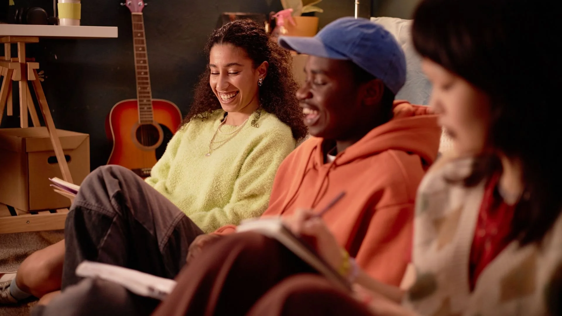 Four young adults sitting on the floor in a cozy room with musical instruments, laughing and taking notes. A guitar is visible in the background.