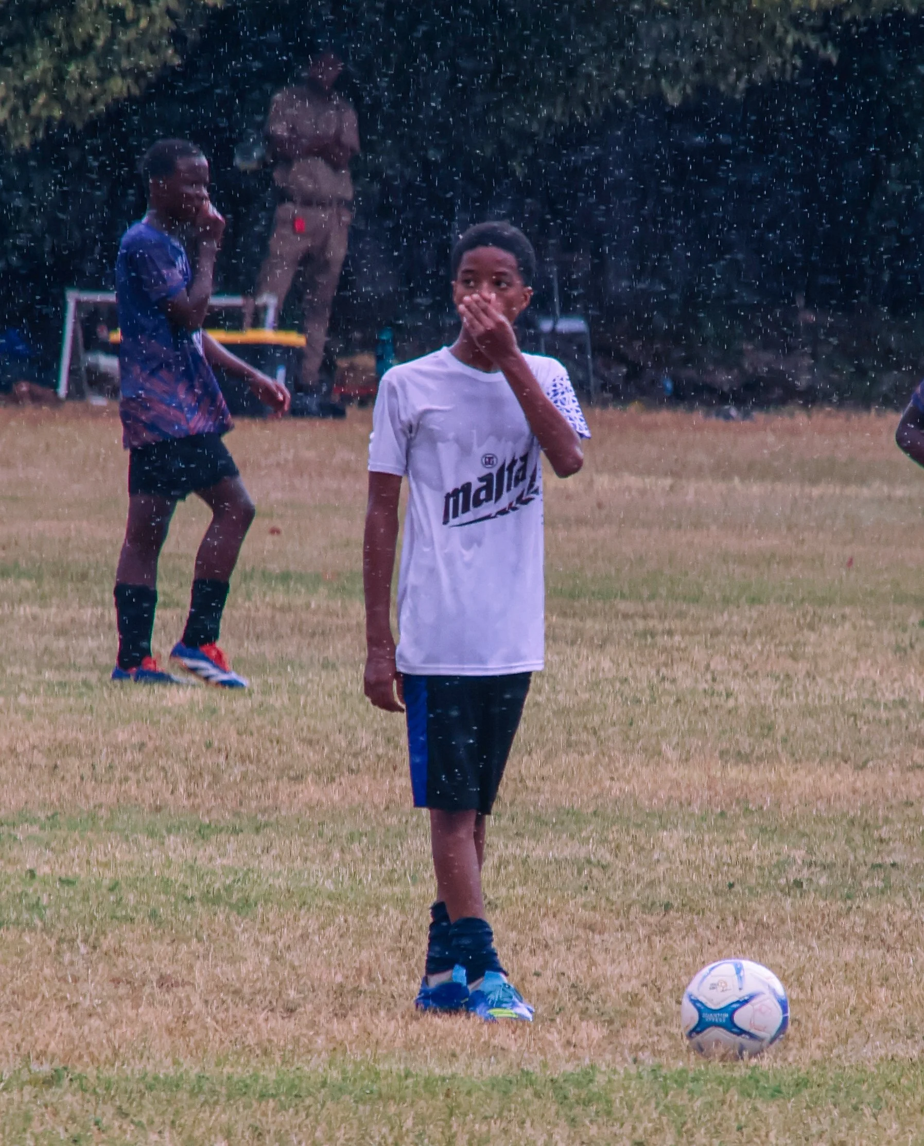 Children playing soccer in a grassy field during rain, with one boy in a white shirt standing near a soccer ball, and other children in the background.