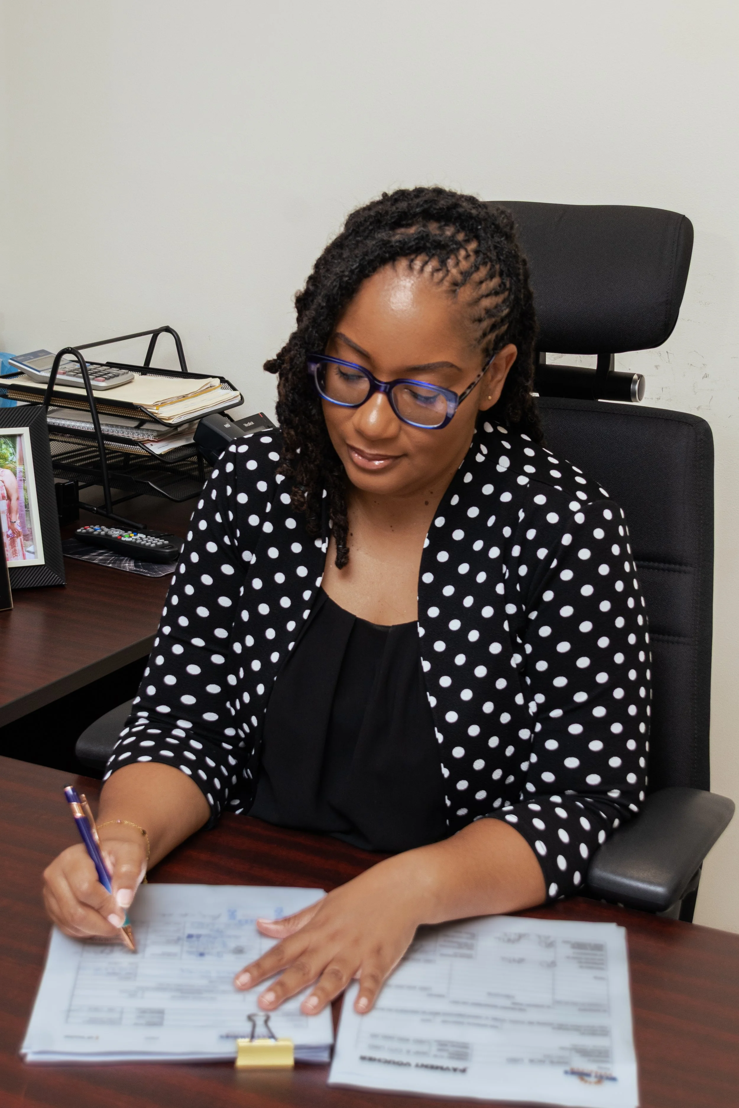 Woman with glasses writing on a document at a desk in an office.