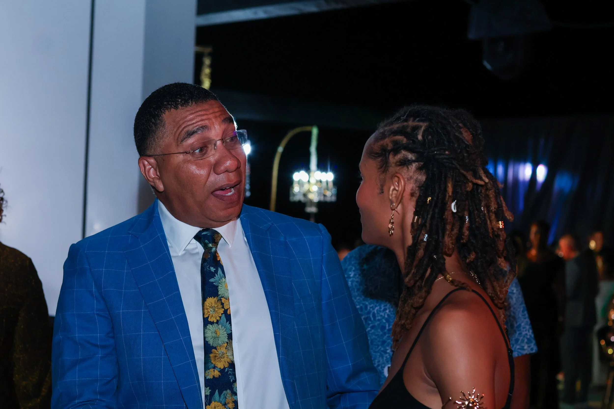 Man in a blue plaid blazer and floral tie talking to a woman with dreadlocks wearing a black dress at a dark indoor event.