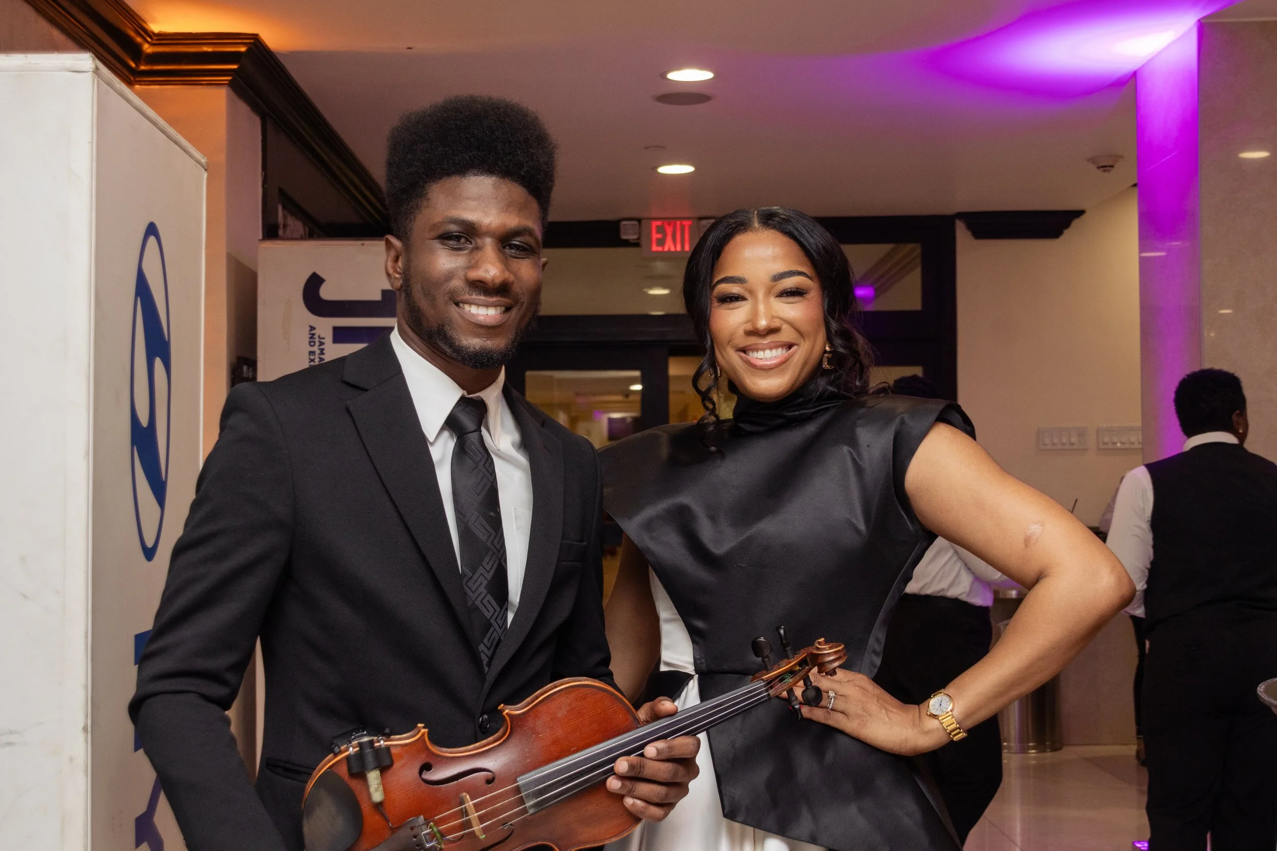 A smiling man and woman posing together at an indoor event, with the man holding a violin. The man is dressed in a black suit and tie, the woman is wearing a black dress. Purple lighting and an exit sign are visible in the background.