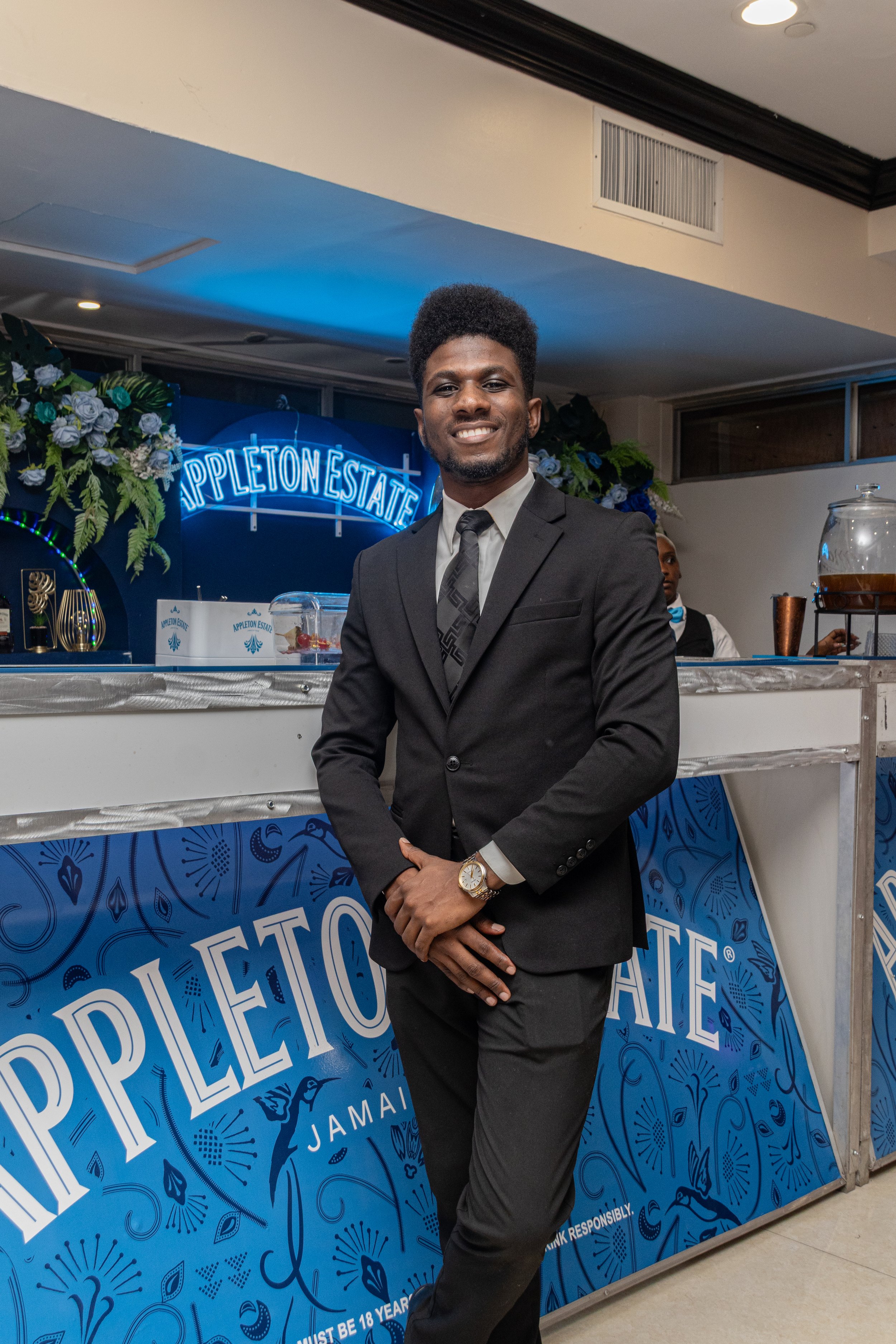 A man in a black suit with a gray tie standing in front of a blue and white promotional display for Appleton Estate rum, smiling at the camera, with a waitress or bartender in the background.