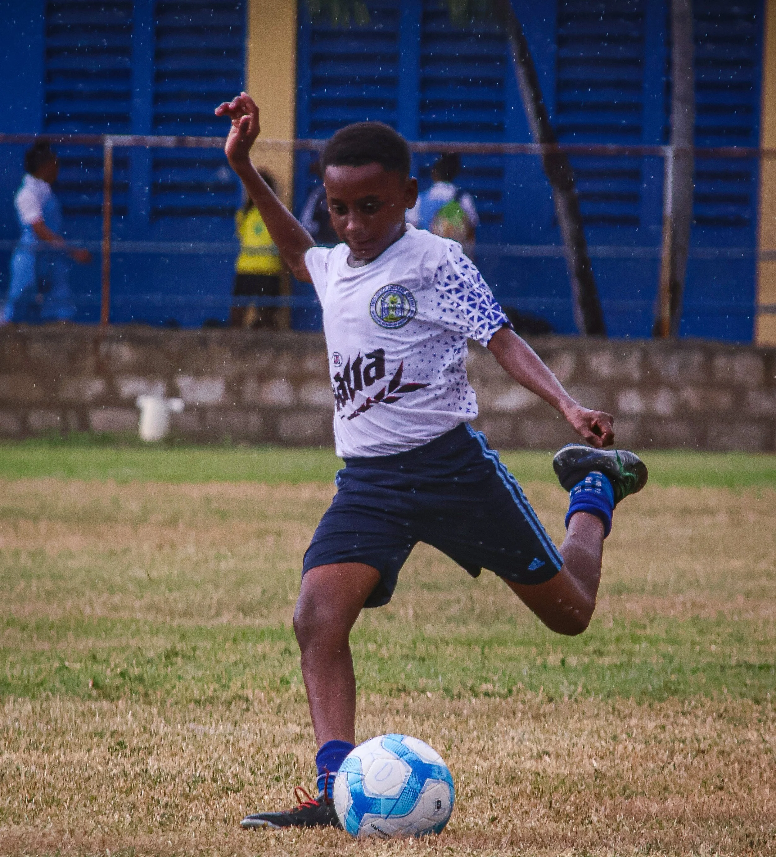 Young boy playing soccer on grass field, kicking a blue and white soccer ball, wearing a white jersey with a logo and black shorts, in evening or indoor setting with blue bleachers in the background.