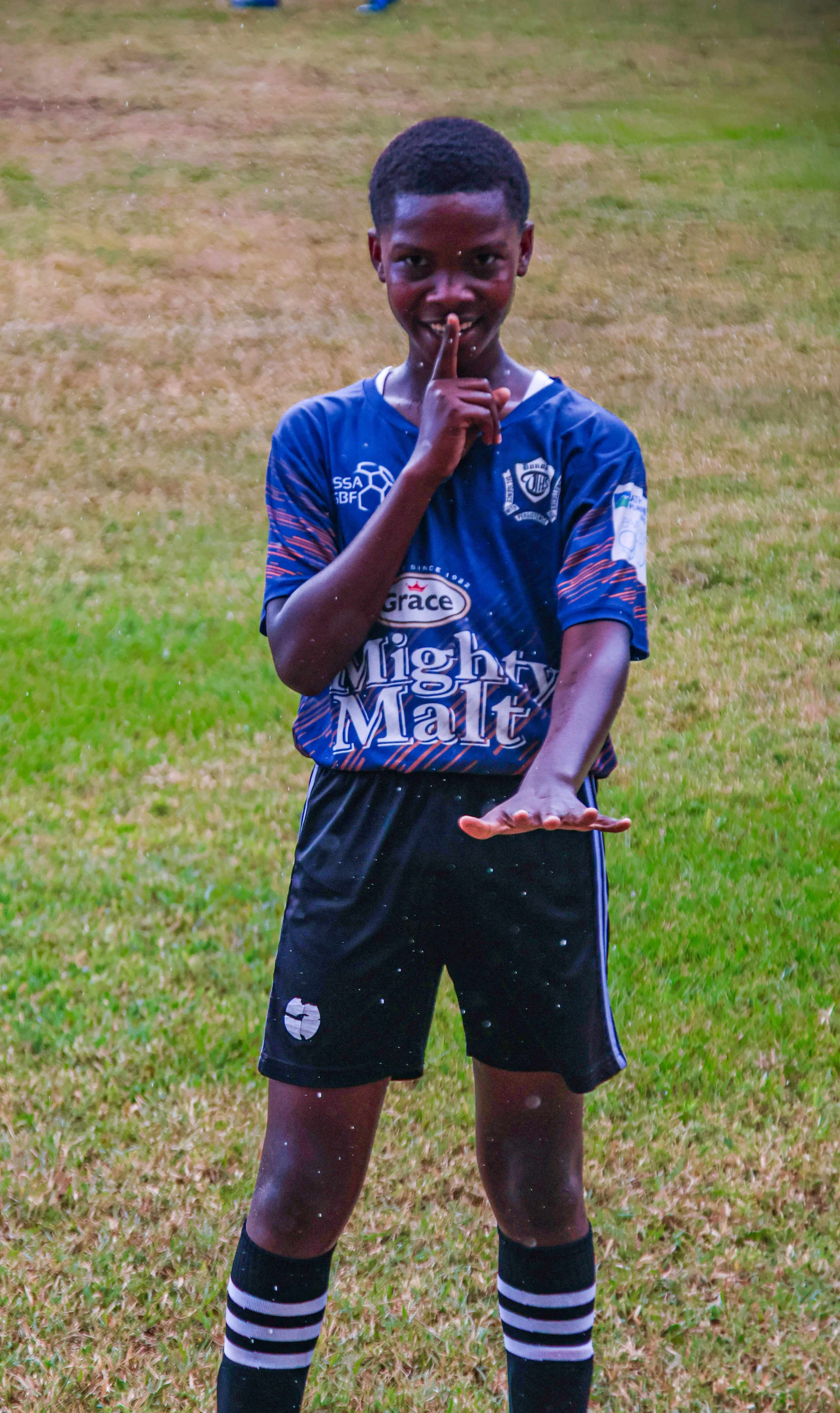 Young boy in football uniform on a field, smiling, holding hand flat below his chin, and making a shushing gesture with his finger.