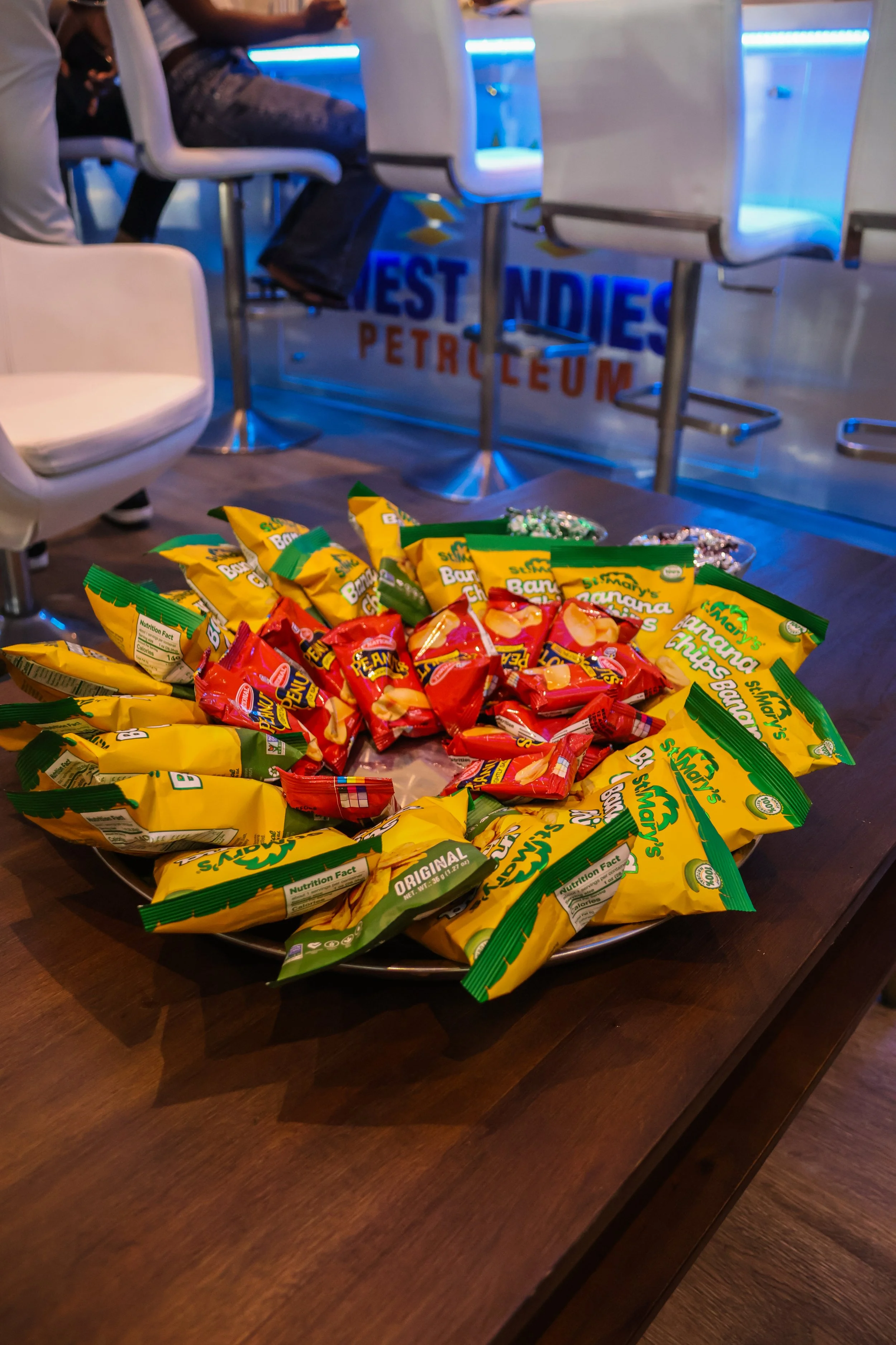 A large metal tray filled with bagged snacks, including banana chips and peanut butter crackers, on a wooden table.