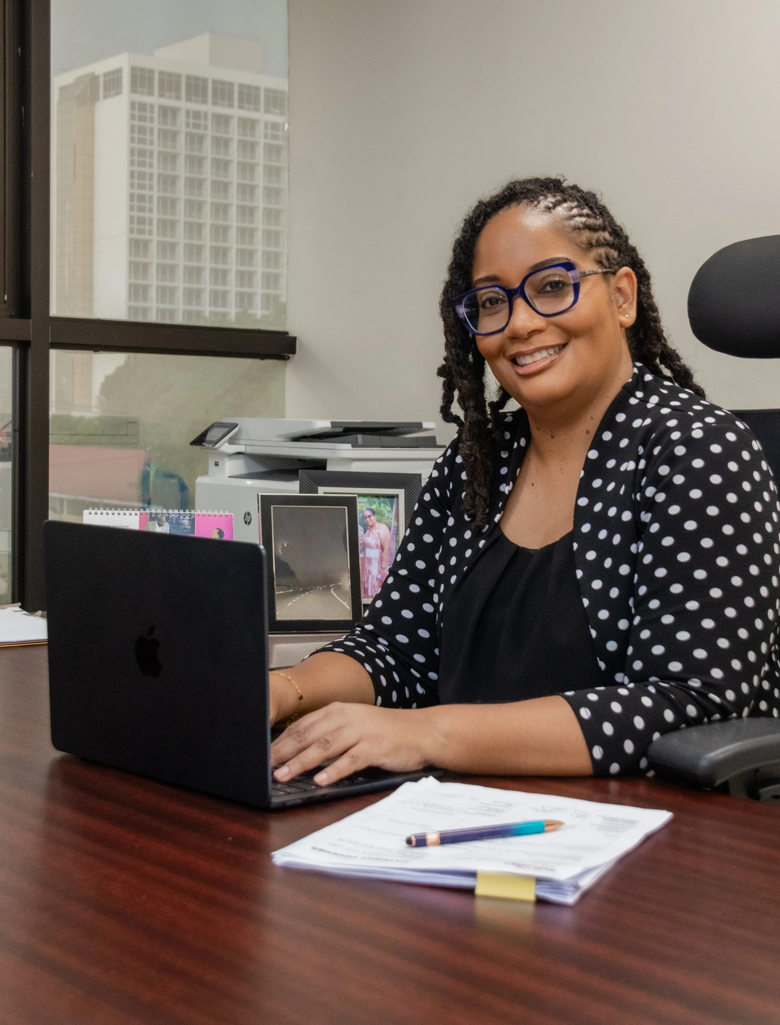 A woman with curly hair, glasses, and a polka dot blazer smiling while sitting at her desk in an office, with a laptop, papers, and a pen in front of her. Behind her are office supplies, a printer, framed photos, and a large window showing a tall bui
