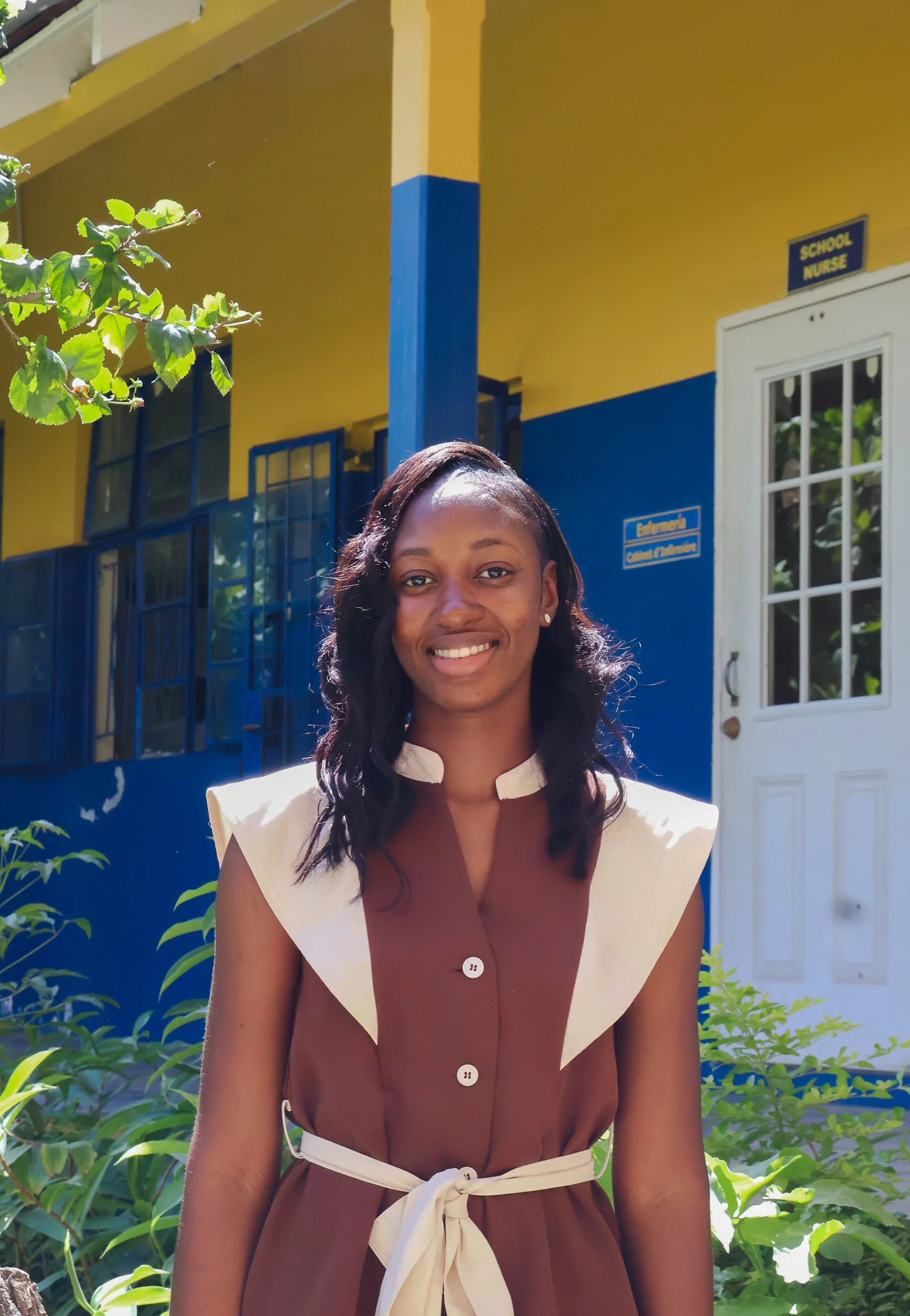 A woman smiling outdoors in front of a yellow and blue building with a sign that reads 'School Nurse' and a window. There are green plants around her.