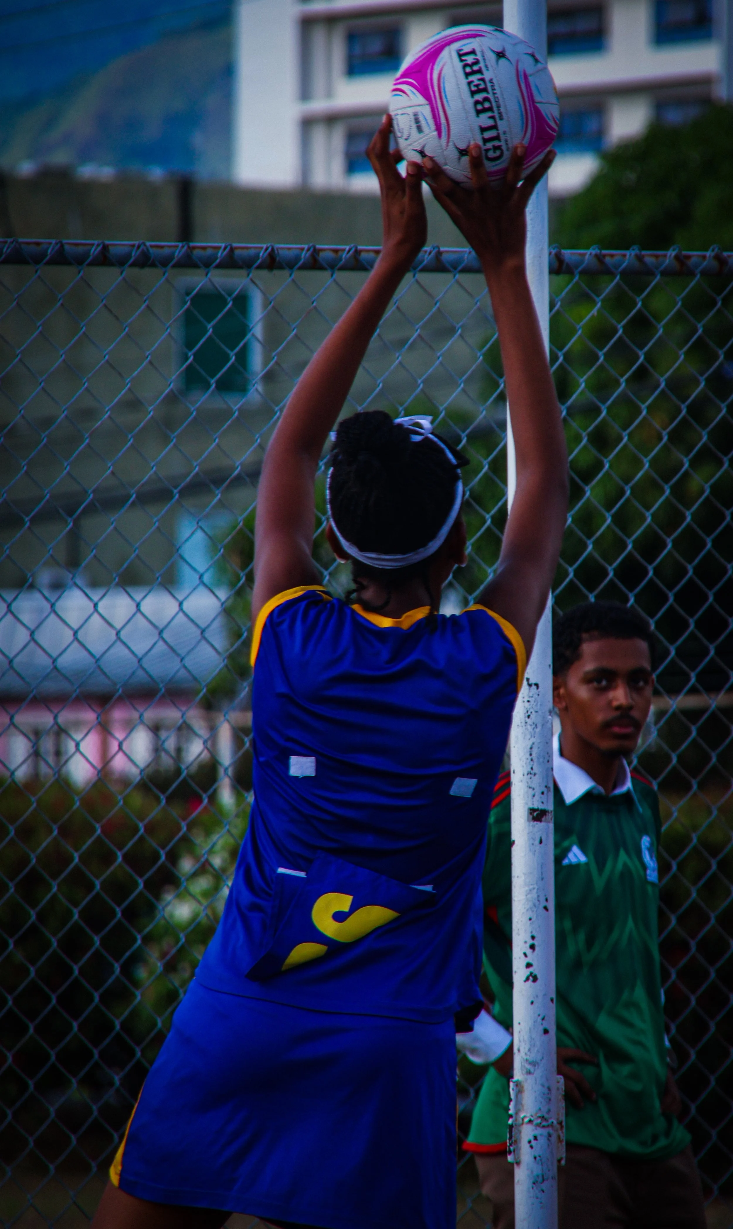 A person in a blue sports jersey is reaching up to hold a white and pink Gilbert brand soccer ball on a soccer goalpost, with a chain-link fence and two other people watching nearby in the background.