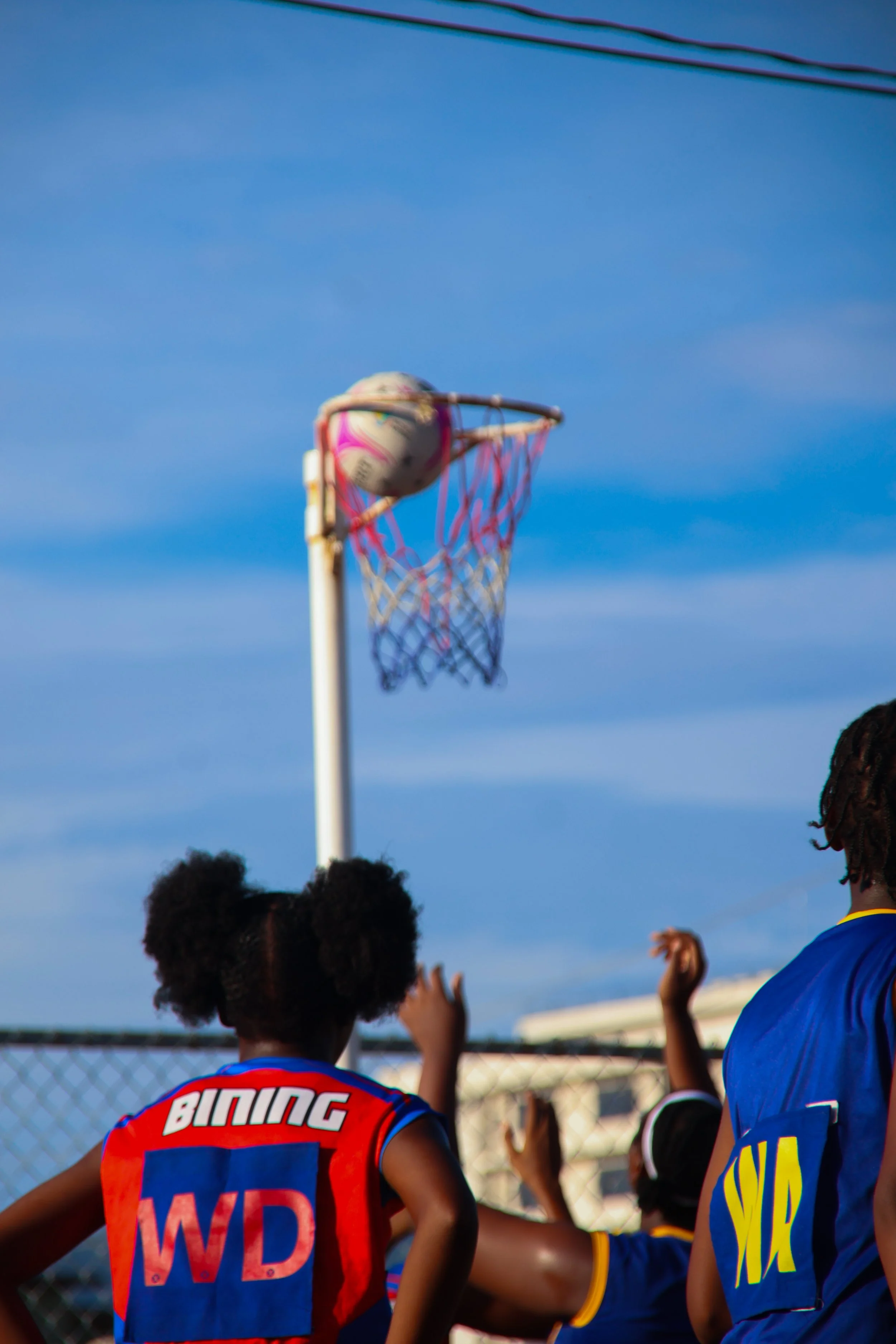 Children playing basketball outdoors near a basketball hoop with a ball in the rim. The kids are wearing colorful jerseys, and some are reaching towards the ball. The background features a blue sky and a chain-link fence.