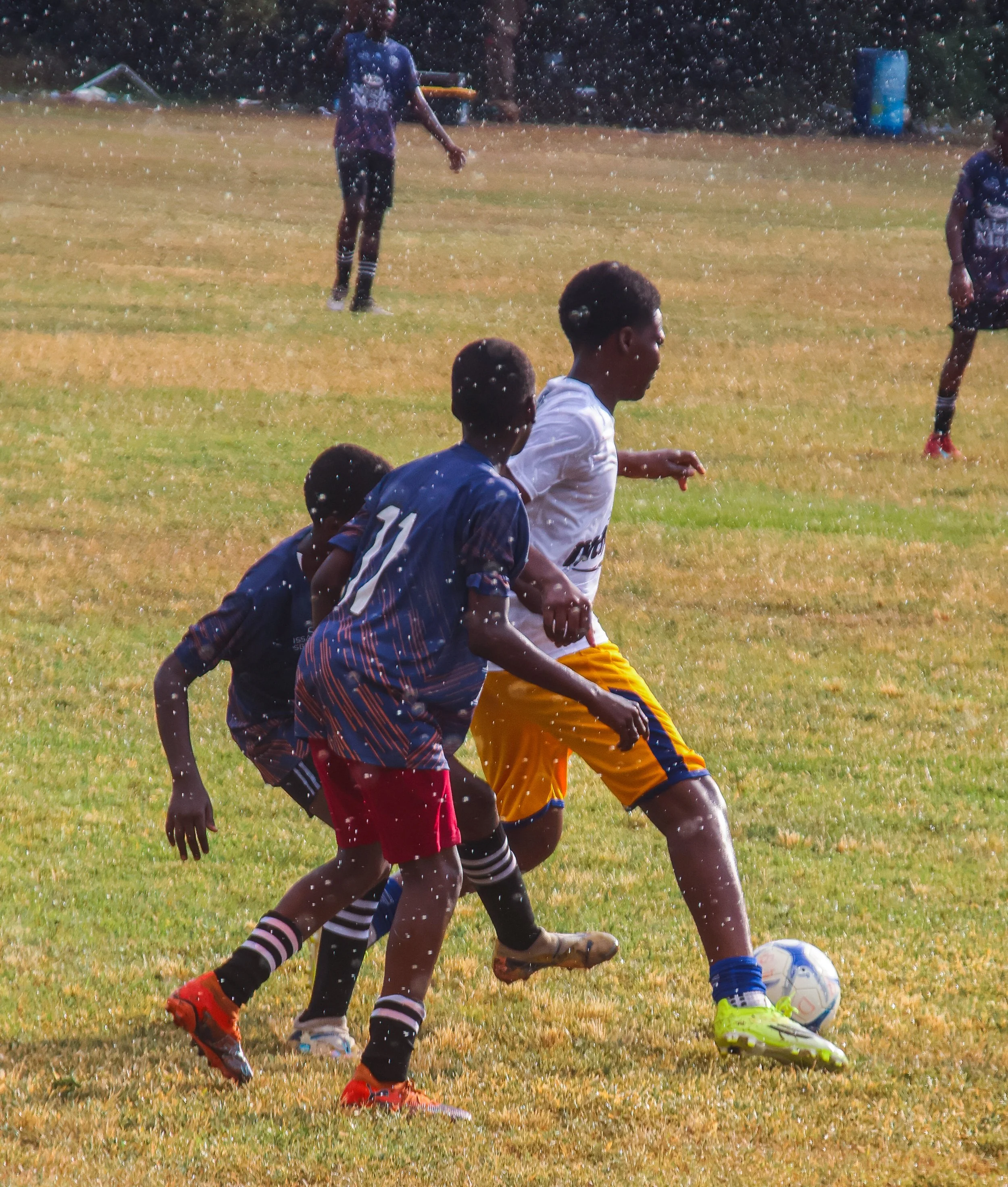 Teenage boys playing soccer on a grassy field during rain, wearing sports uniforms and boots, with one boy in a white shirt and yellow shorts kicking the ball.