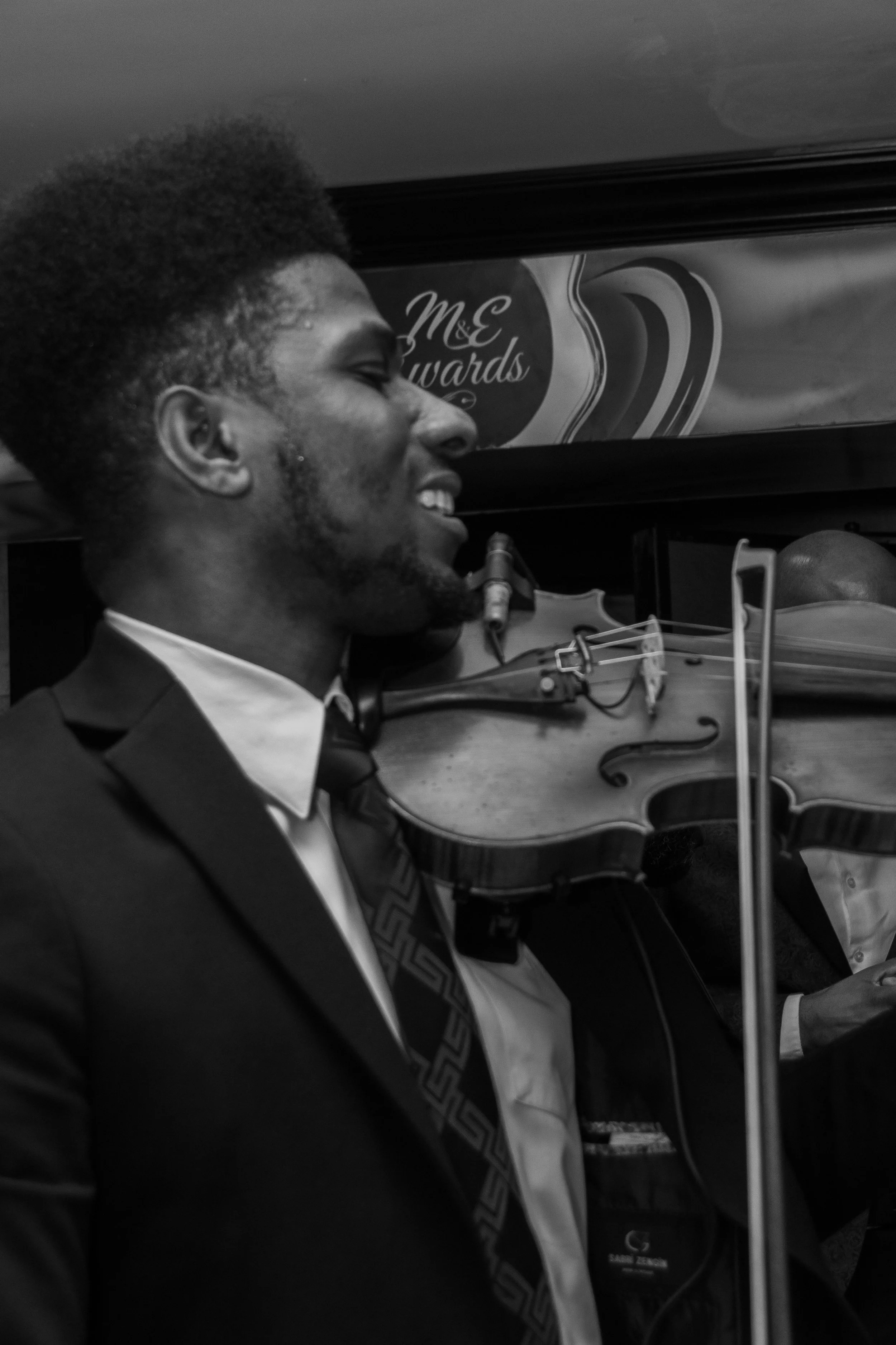 A man in a suit and tie holding a musical instrument, possibly a violin, appears to be smiling or singing, with a banner in the background that reads 'M&E Awards'.