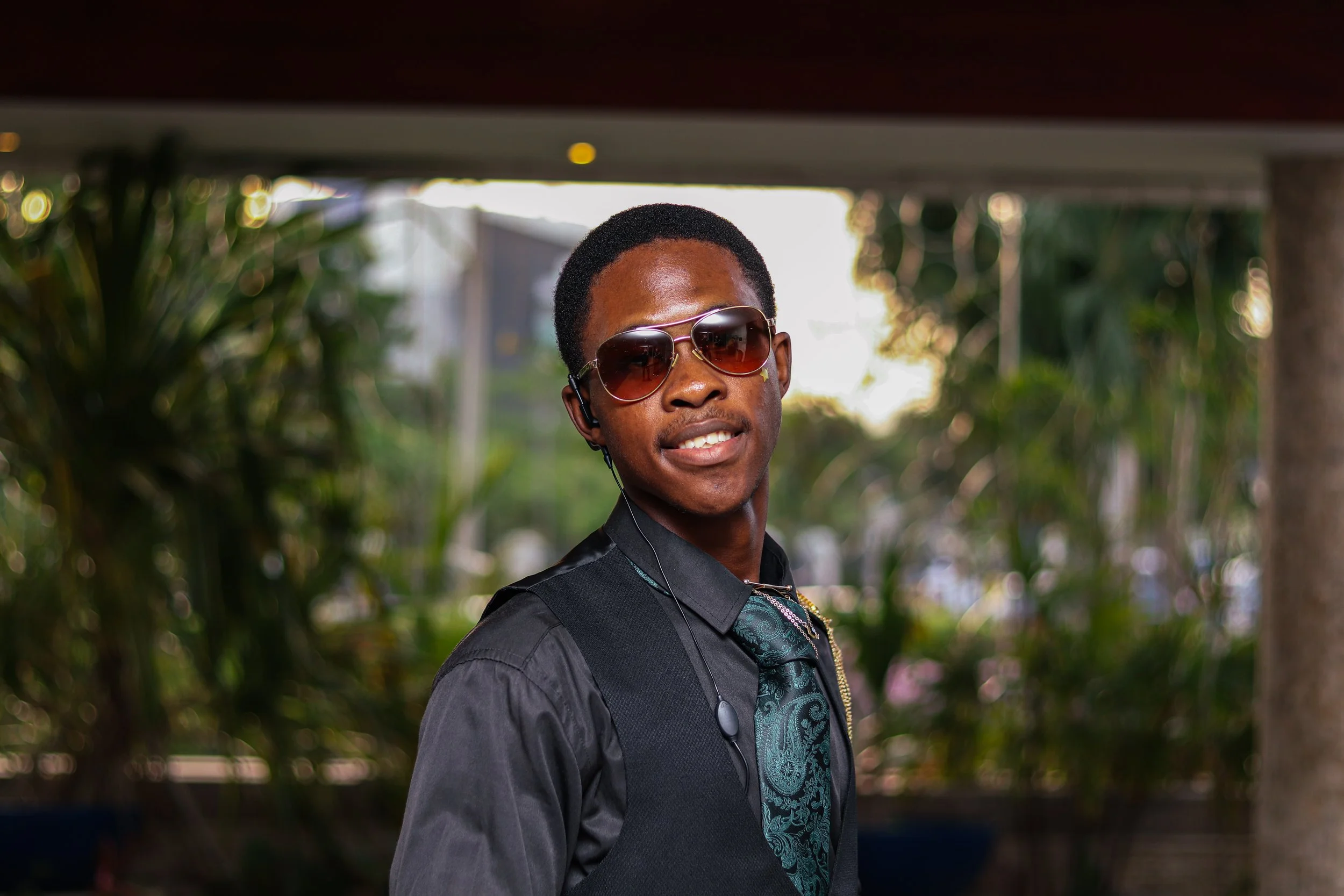 A young man with dark skin wearing sunglasses, a dark shirt, and a patterned tie, standing outdoors with blurred green foliage in the background.