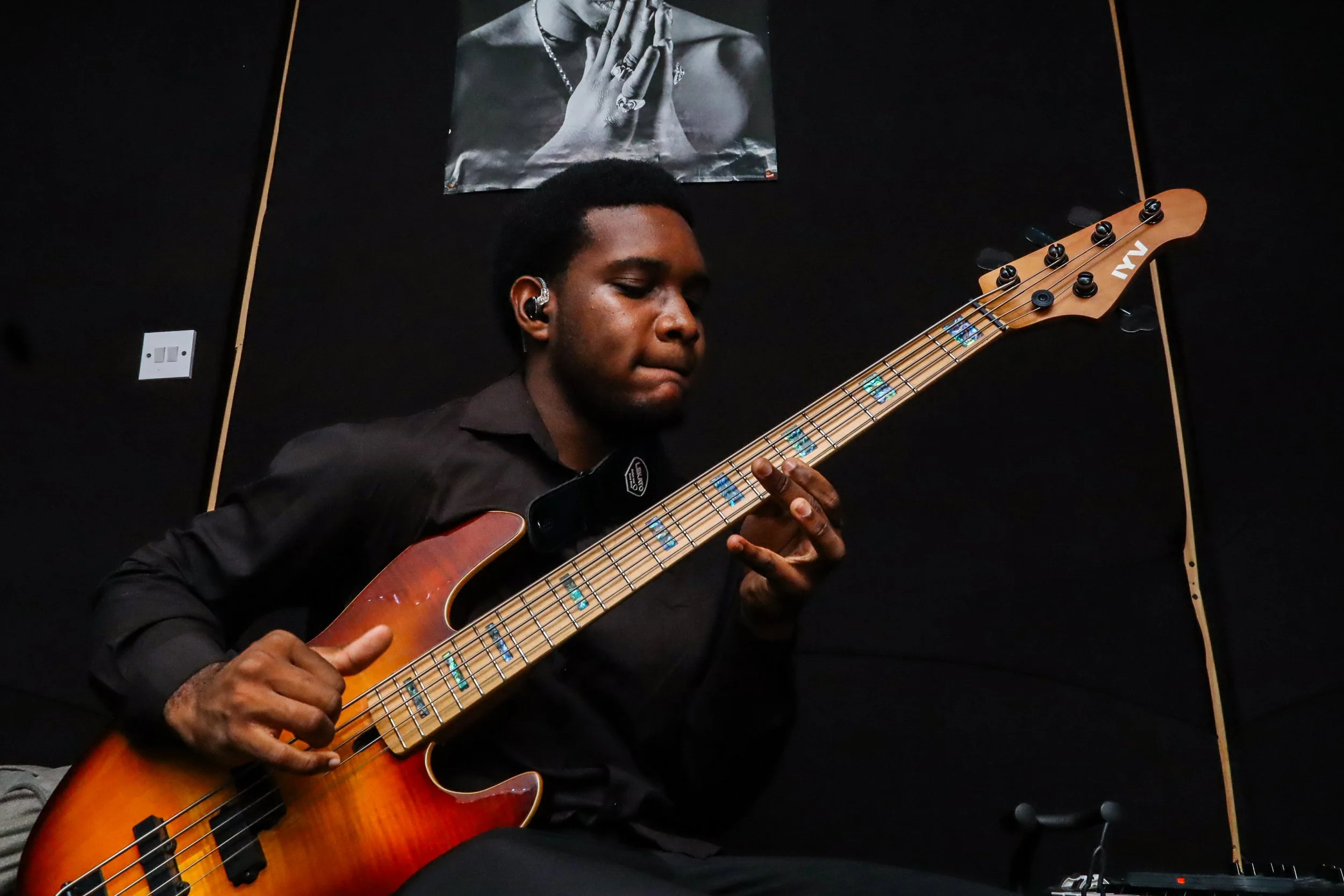 A young man playing an orange and brown electric bass guitar with blue inlays, wearing a black shirt, in a dark room with a black wall and a poster of a person with hands clasped in prayer behind him.