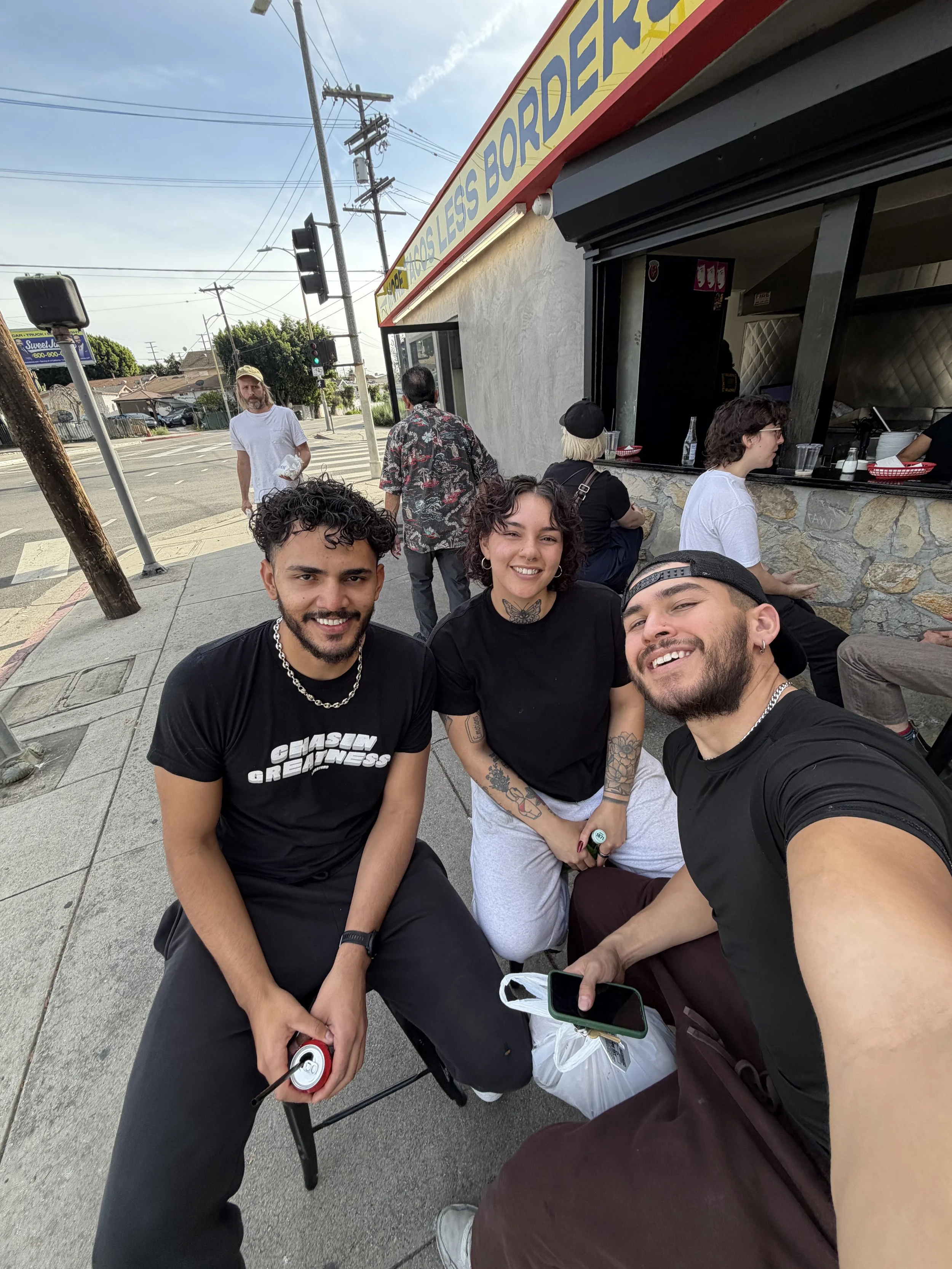 Three young adults sitting outside a restaurant taking a selfie, with a woman smiling, a man in black with a chain necklace, and a man in a backwards cap in the foreground, while other people are visible in the background near the restaurant window, with an urban street scene behind them.