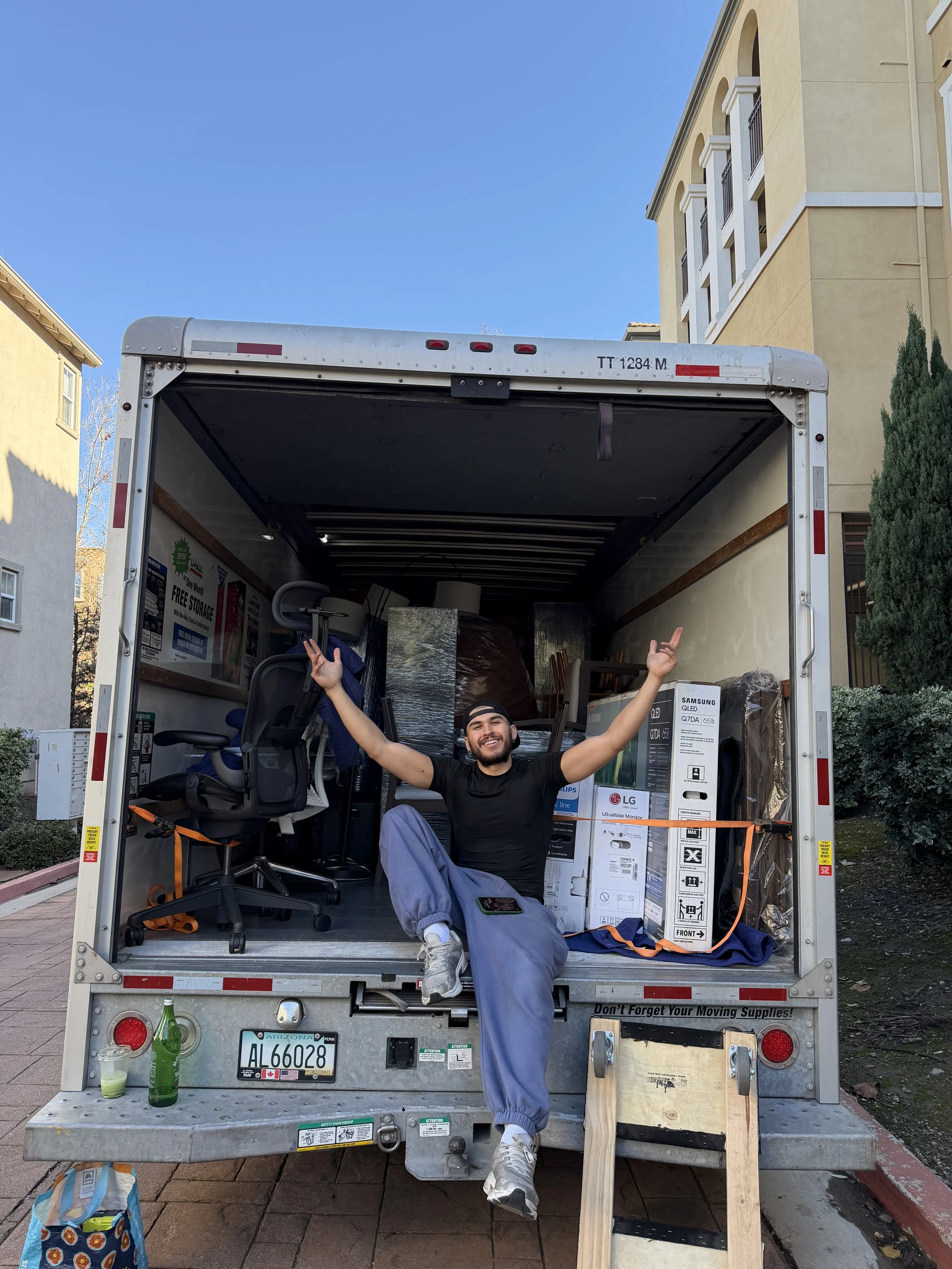 A smiling man with a beard and cap sitting inside an open moving truck, with arms raised. The truck is filled with furniture and boxes, including electronics and a chair. Outside the truck are a green bottle, a drink, a bag, and a wooden plank, in a residential area with multi-story buildings and trees.