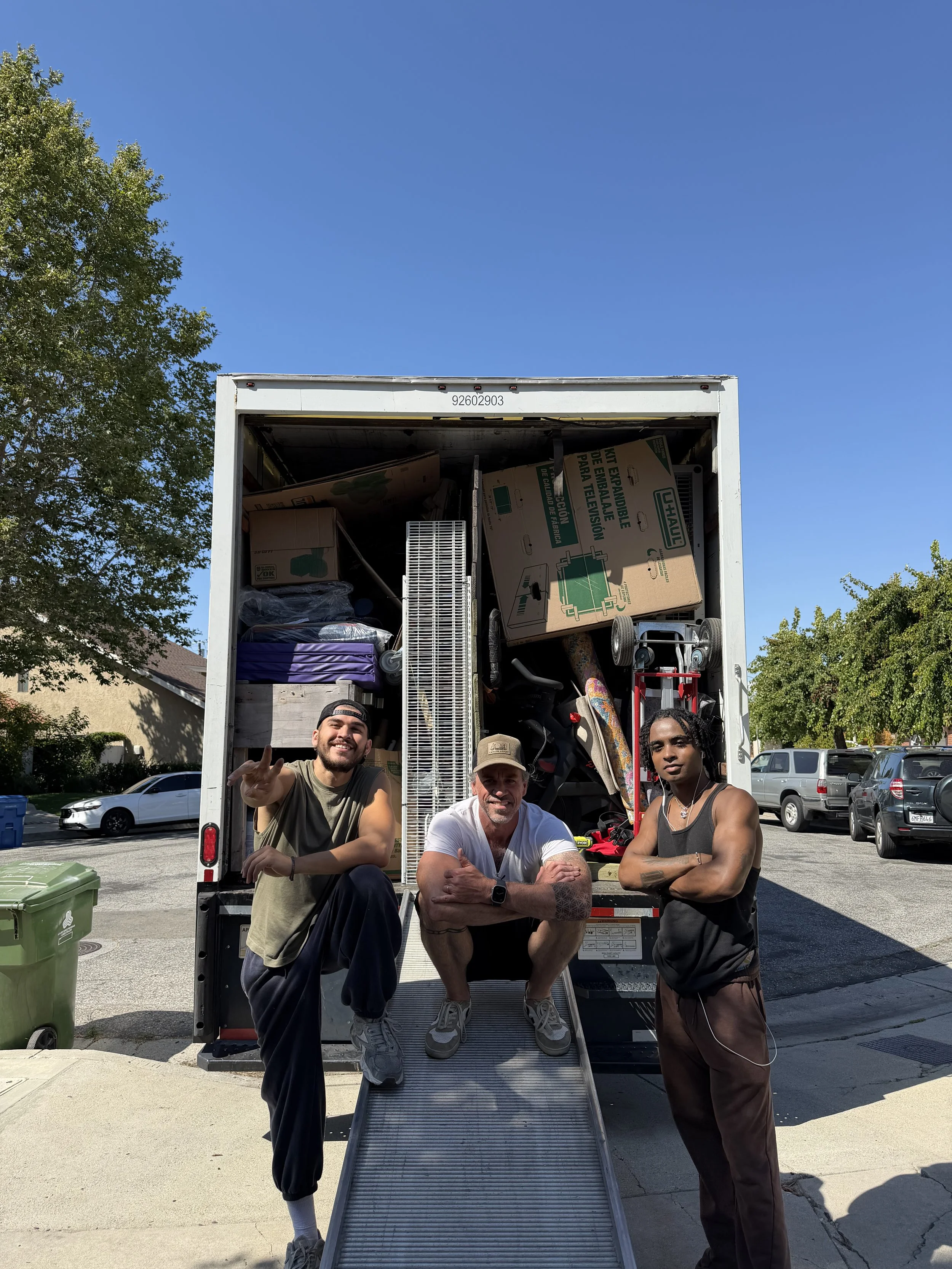 Three men stand in front of an open moving truck filled with boxes and furniture. They are outdoors on a sunny day, with blue skies and trees in the background.