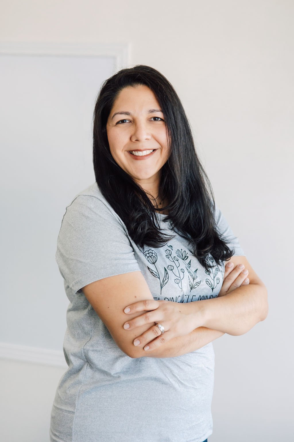 A woman with long black hair, smiling and crossing her arms, wearing a light gray T-shirt with floral design, standing against a plain white wall.