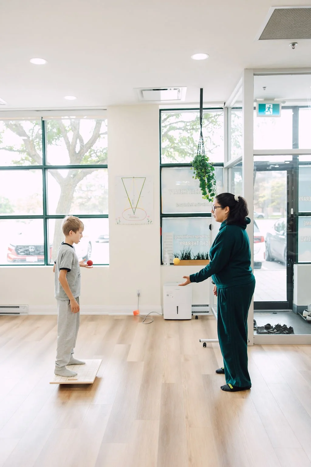A boy standing on a wooden platform in a therapy or exercise room, facing a woman who appears to be a therapist or trainer, holding a small red ball in her hand. The room has large windows, indoor plants, and some therapy or exercise equipment.