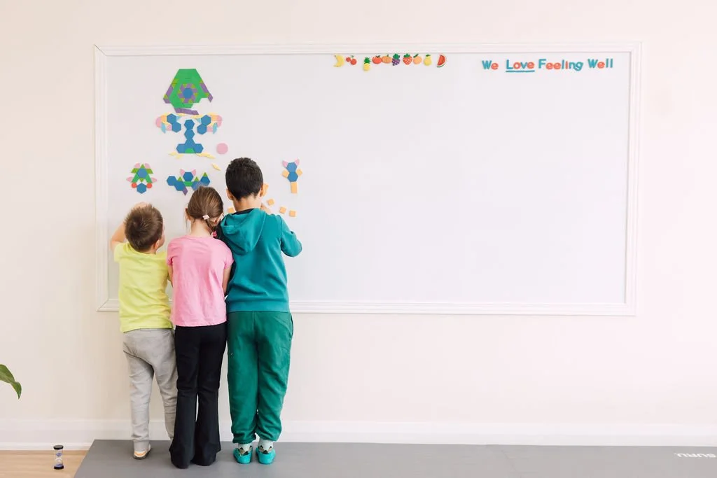 Three children standing in front of a whiteboard, engaging with colorful geometric shapes and stickers.