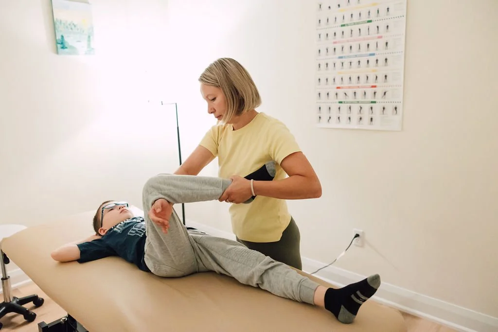 A physical therapist assisting a young boy with a leg exercise on a treatment table in a clinic room.