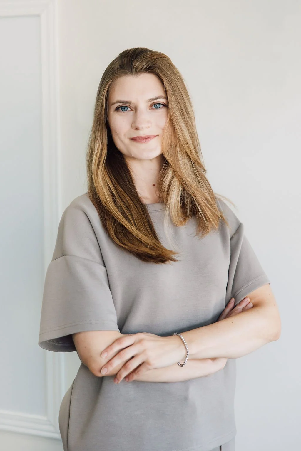 A woman with long, light brown hair and blue eyes crossing her arms and smiling softly, standing against a neutral background.