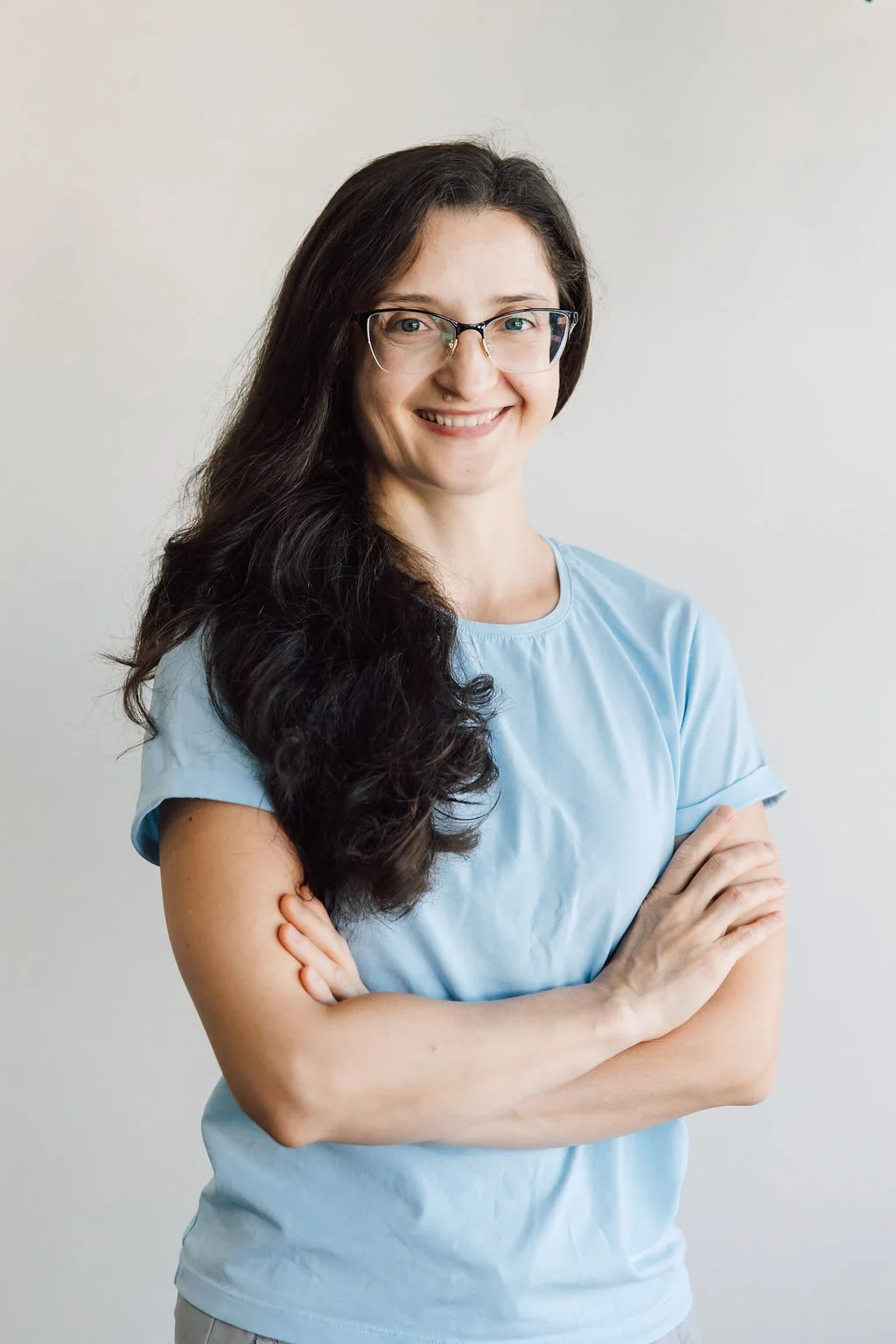 Smiling woman with glasses and long dark hair, wearing a light blue shirt, standing with arms crossed against a plain white background.