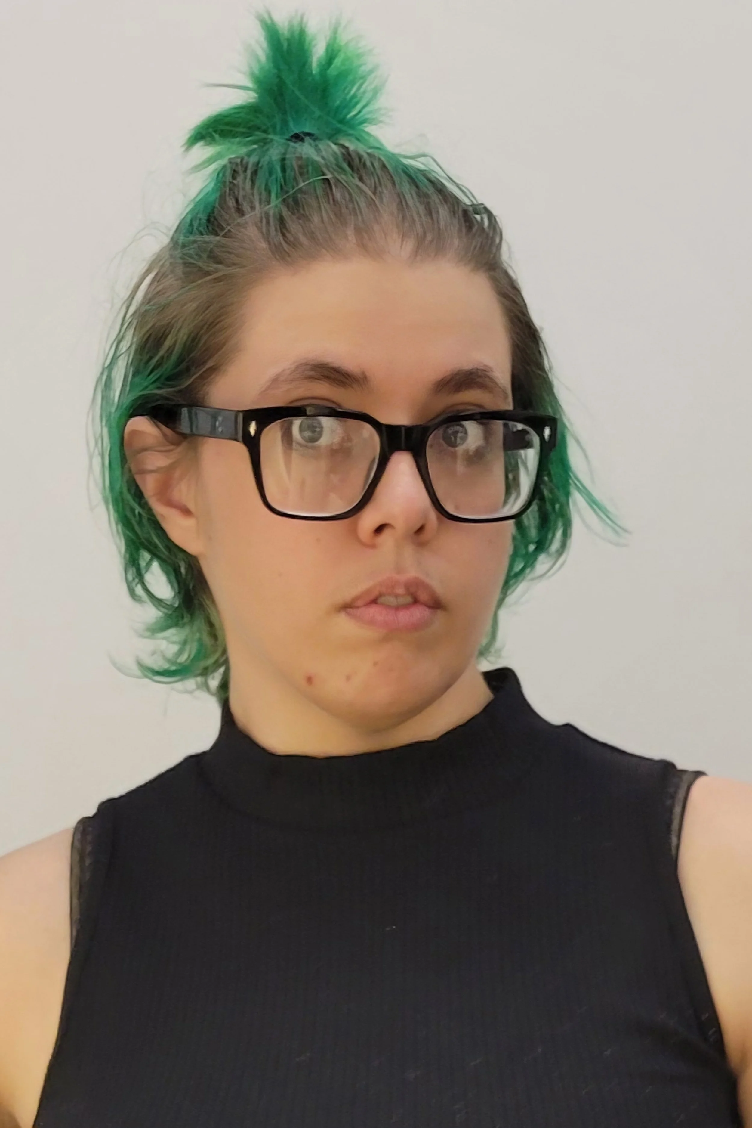 A woman with green hair styled in a high bun, wearing black glasses and a black sleeveless top, looking slightly to the side against a plain light-colored background.