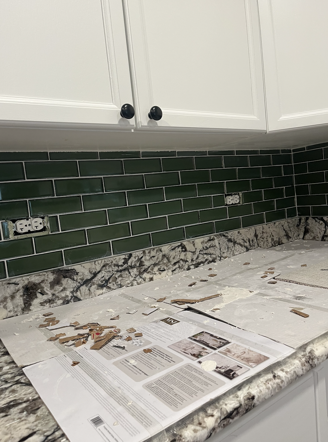 Kitchen countertop with green subway tile backsplash, white cabinets above, and a newspaper with cut-up pieces on the surface.