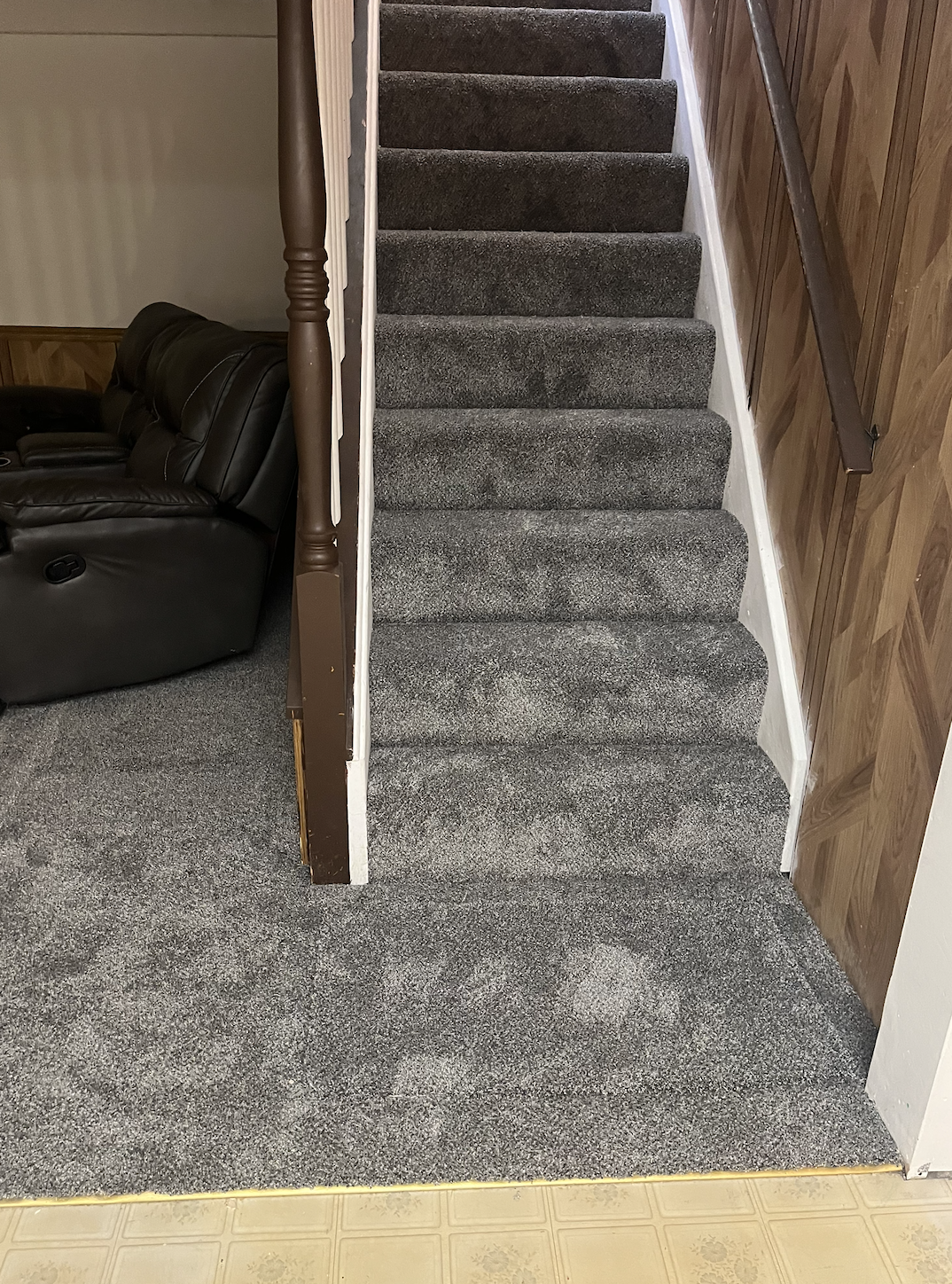 Indoor scene featuring a carpeted staircase with dark gray carpeting, a wooden handrail on the right, a beige wall on the left, and part of a dark brown recliner sofa on the left side of the image.