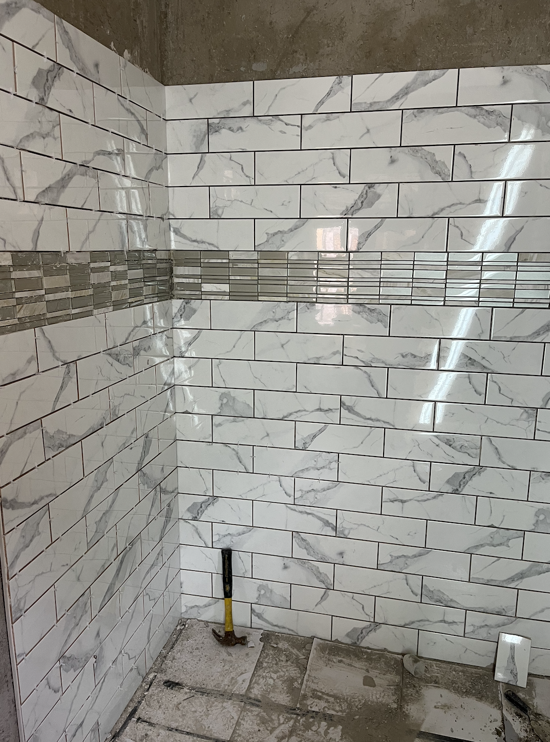 A corner of a bathroom with white marble tiles and a horizontal strip of gray mosaic tiles on the wall, with construction tools on the floor.