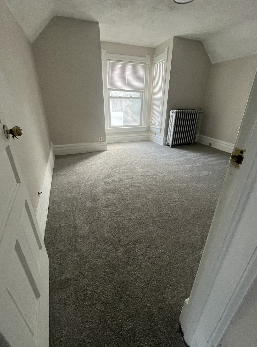 Empty room with carpeted floor, two windows with blinds, a radiator, and plain light-colored walls.