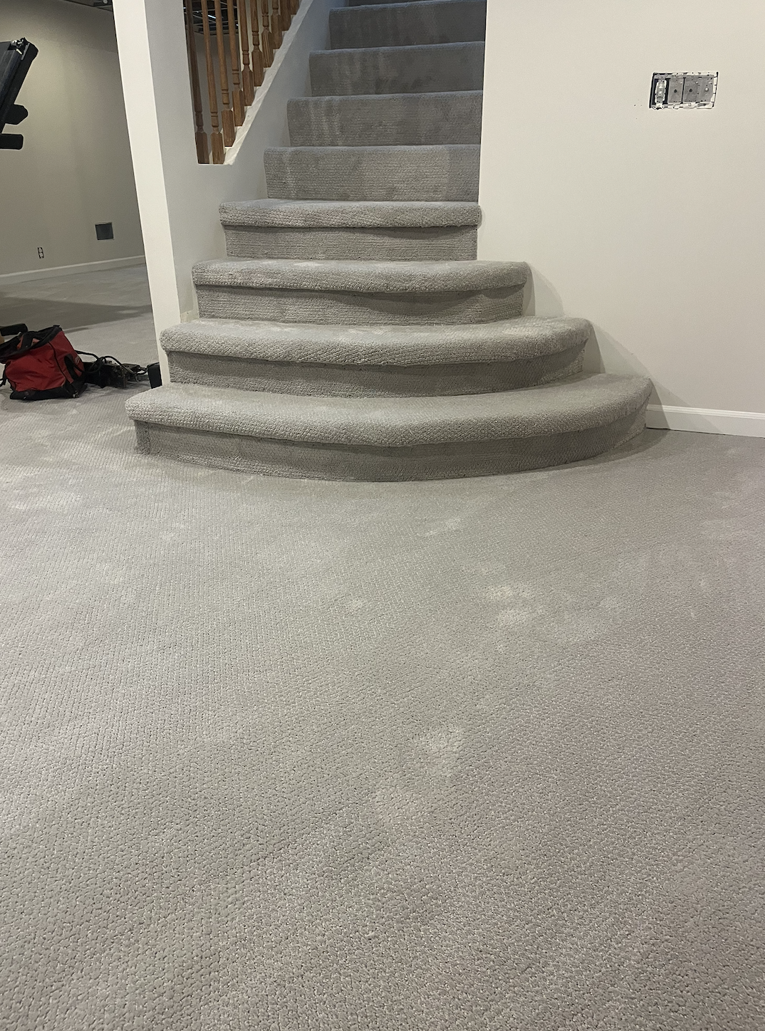 The image shows a beige carpeted staircase with rounded edges, leading upward, with a wooden railing on the left side. The continuing carpeted floor is visible at the bottom, with a red and black bag placed on the left side near the wall. An electric