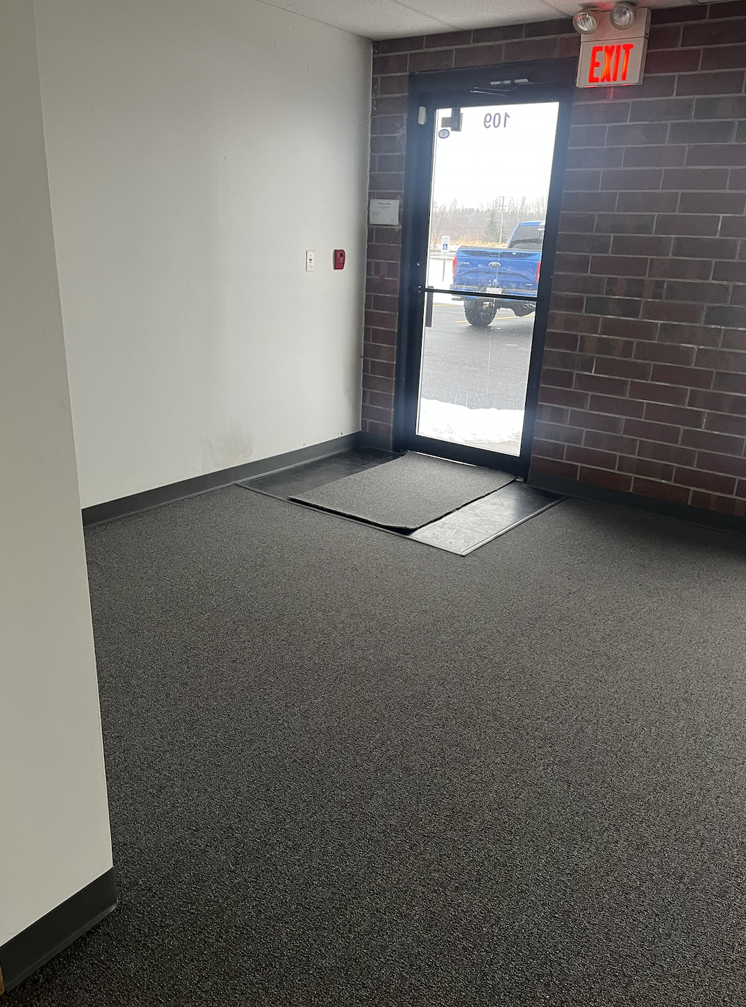 Inside view of a building entrance with a glass door, red EXIT sign, carpeted floor, and outside parking lot with a blue truck.