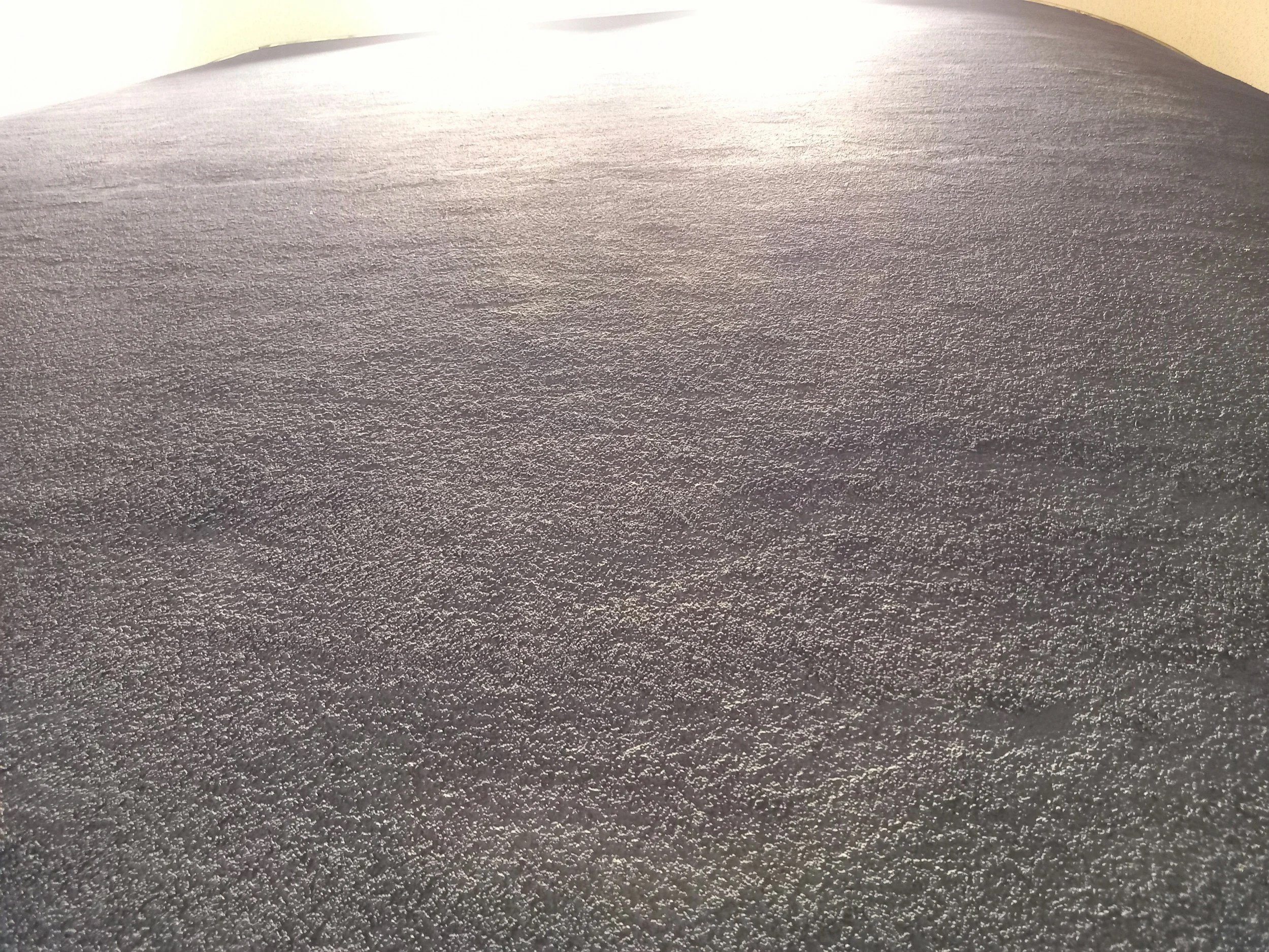 Close-up of a textured, dark-colored surface, possibly a mat or carpet, with a slightly uneven edge and a light-colored wall or surface in the background.
