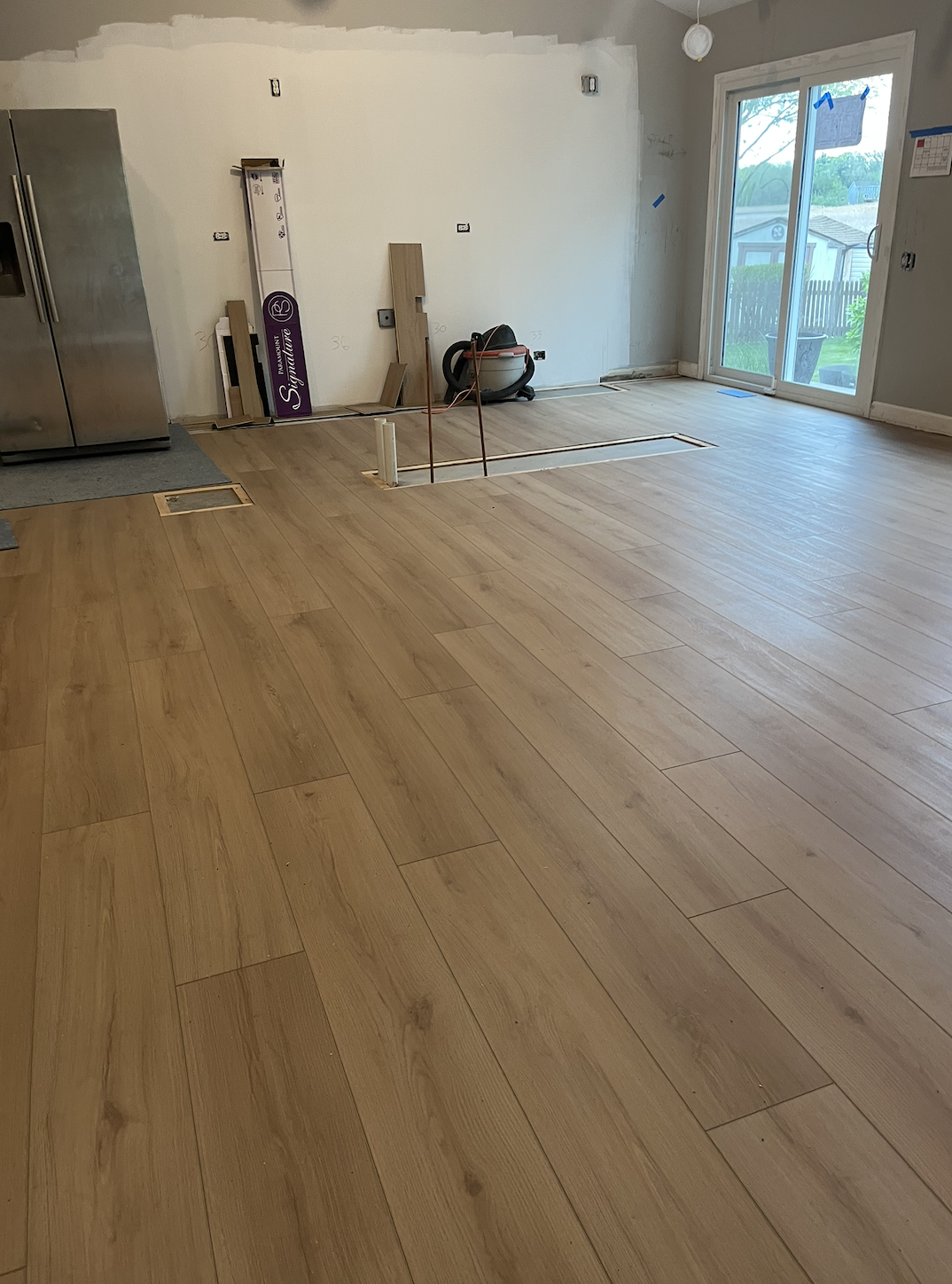 A room under renovation with new hardwood flooring partially installed, tools and materials visible including a vacuum, wood planks, and construction markings on the wall. A sliding glass door leads outside.