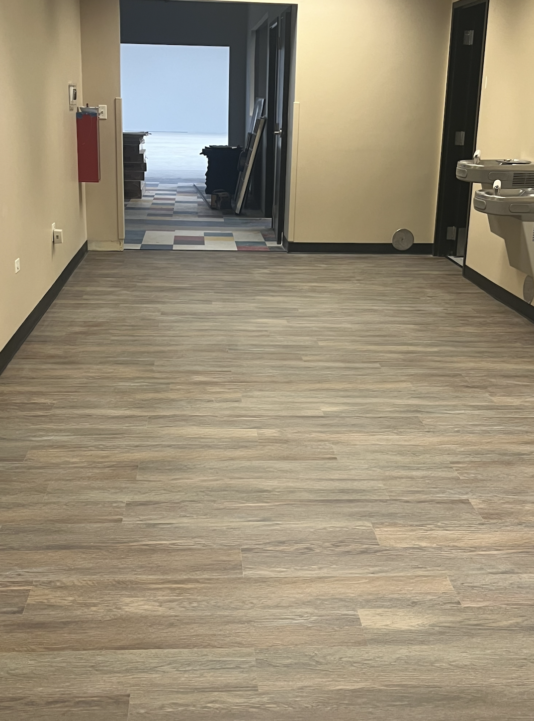 Empty room with light wood flooring, beige walls, and black trim, leading to a hallway with a patterned carpet and construction materials at the end.