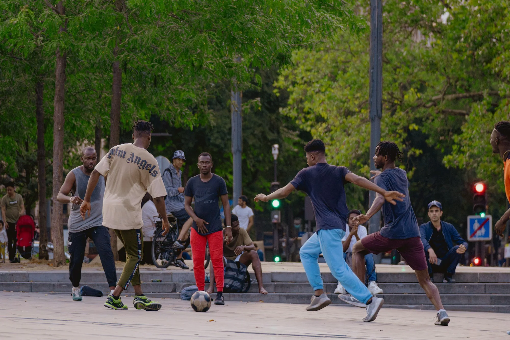 Adults playing a pickup game of street soccer on pavement under the trees