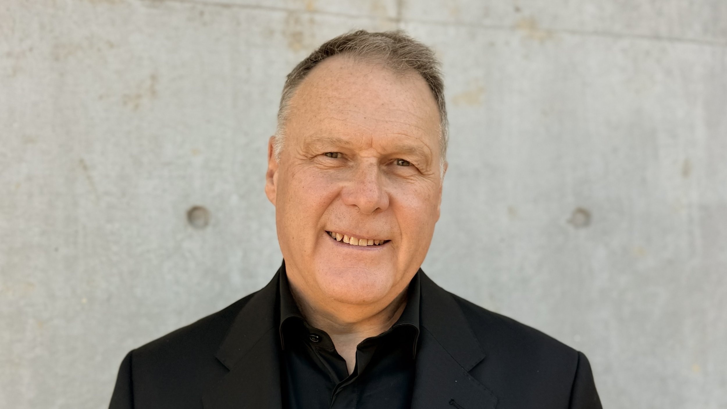 A middle-aged man with short, light hair and a friendly smile, wearing a black suit and black shirt, standing against a plain concrete wall.
