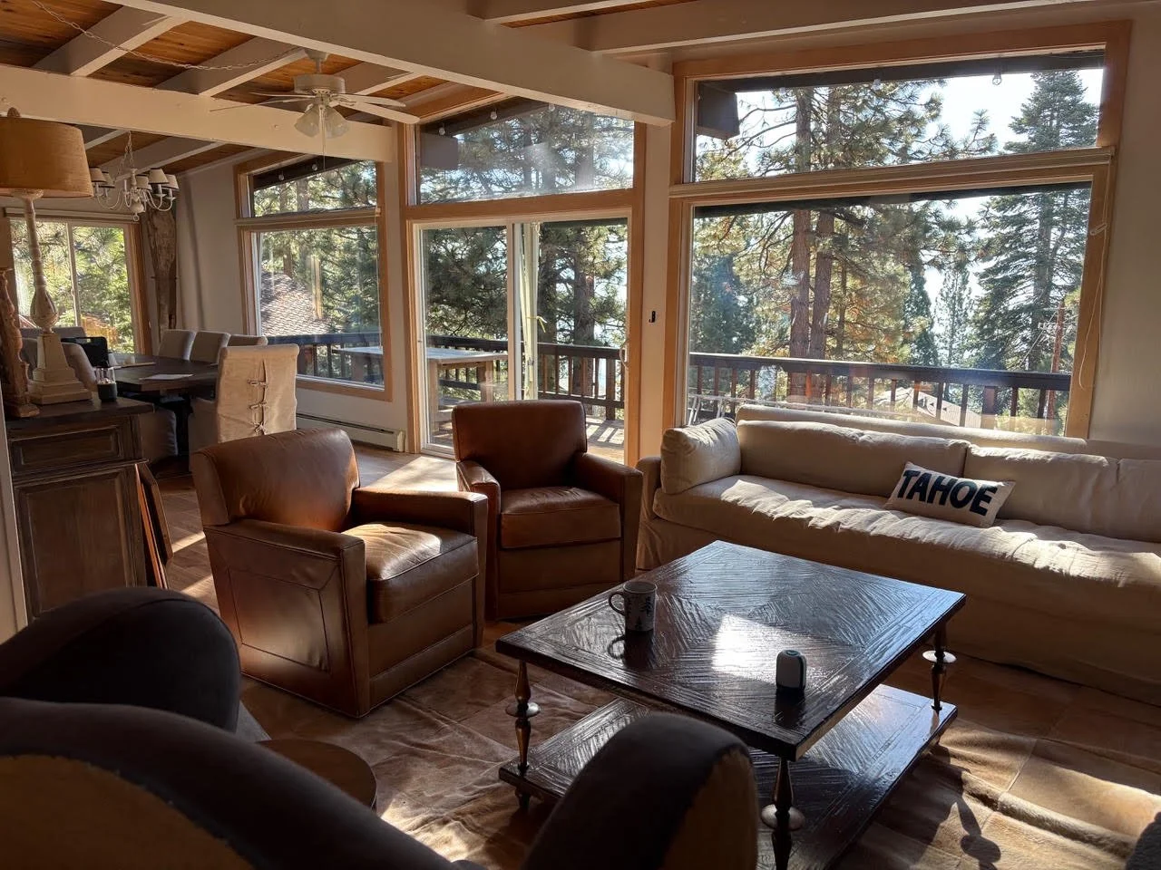 Living room with large windows showing a forest view, and a dining area in the background with a chandelier.