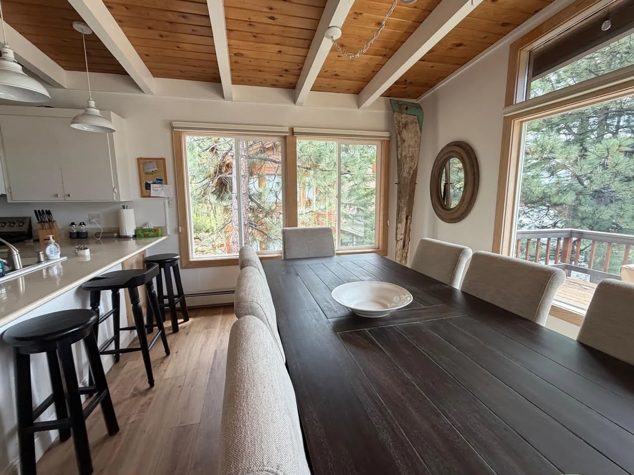 Dining room with a dark wooden table surrounded by beige upholstered chairs, large windows showing trees outside, a white kitchen counter with black stools, and a wooden ceiling with beams.