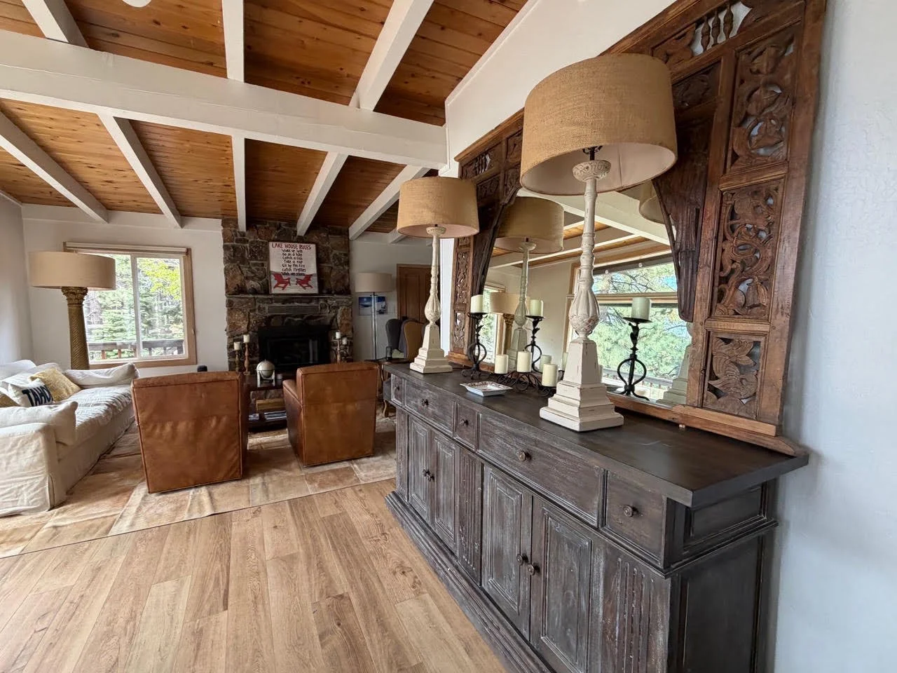 A large living and dining room with wooden beamed ceiling, stone fireplace, with windows overlooking trees and the lake.
